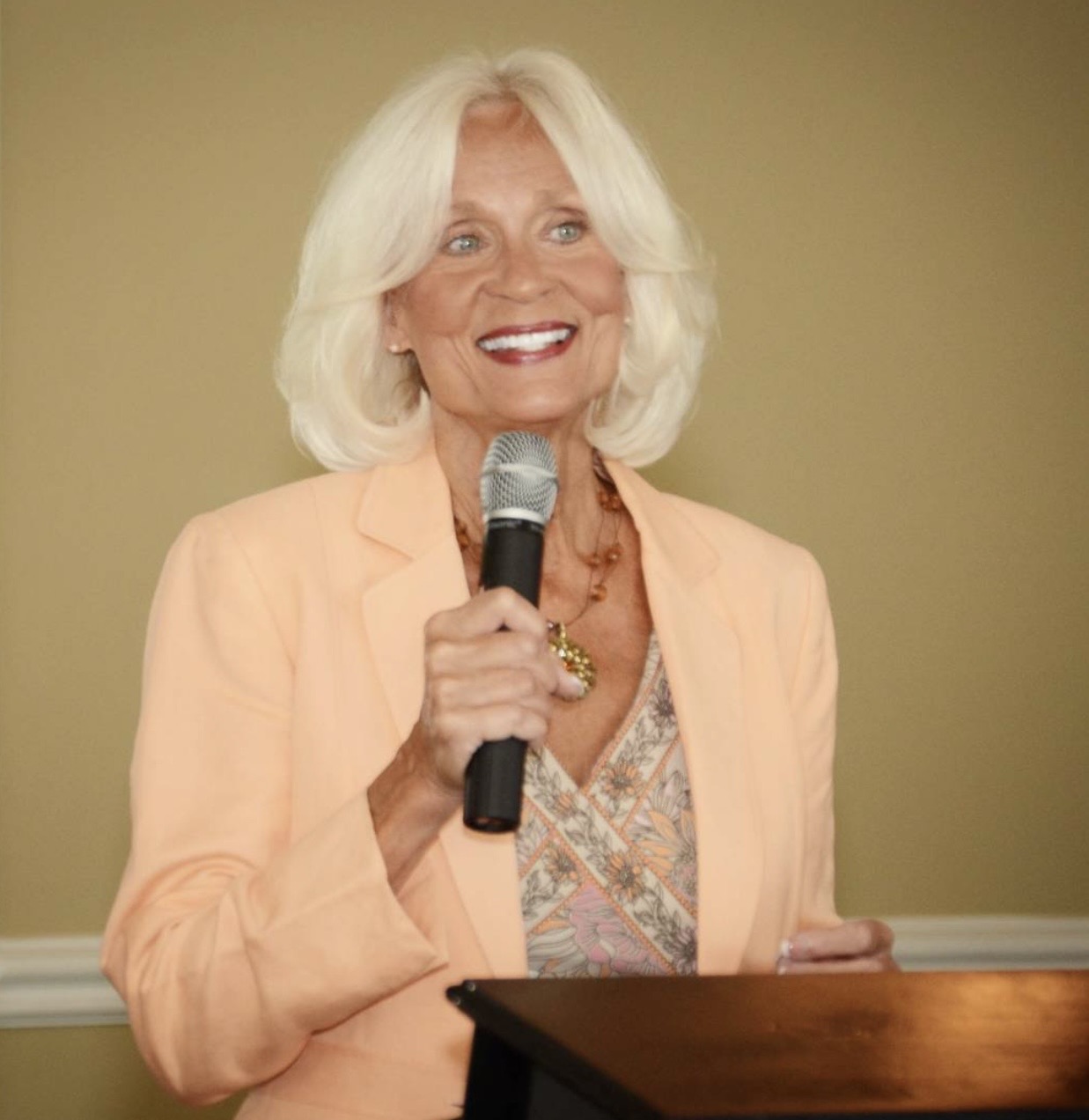 Older woman with white hair smiling, holding a microphone, wearing a peach blazer and patterned blouse, standing at a podium.