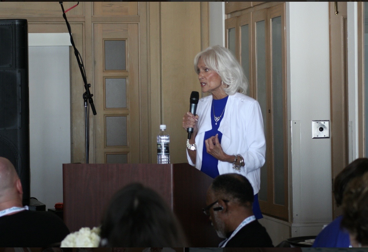 Woman with white hair speaking into a microphone at a podium during a presentation or event.
