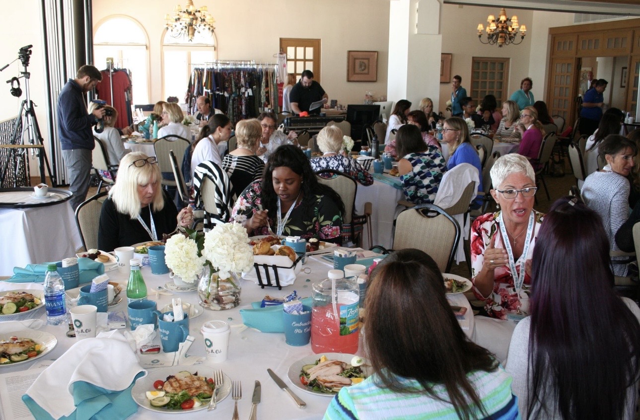 People seated at round tables in a large room, eating and socializing during a gathering or event.