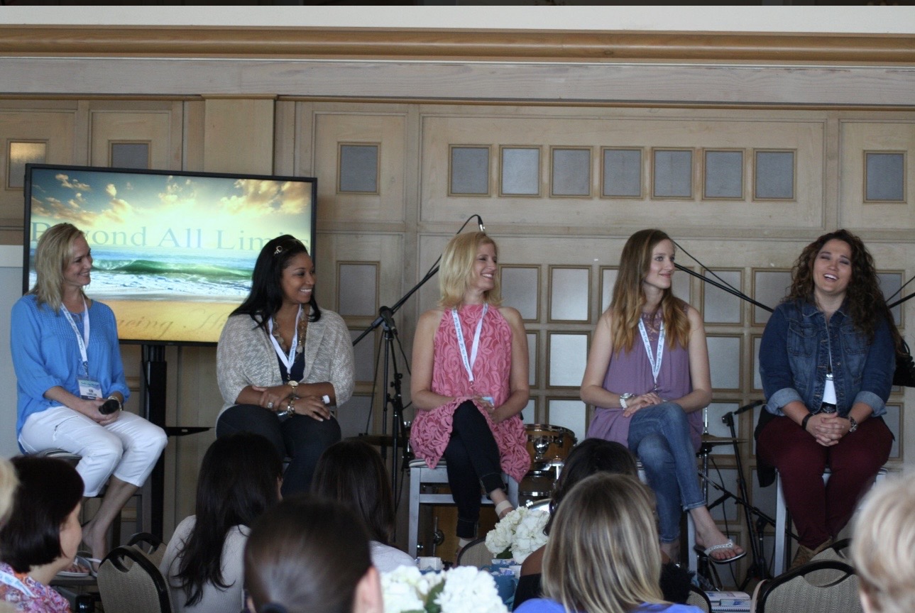 Five women sit on chairs on stage, with a screen behind them displaying text and graphics.