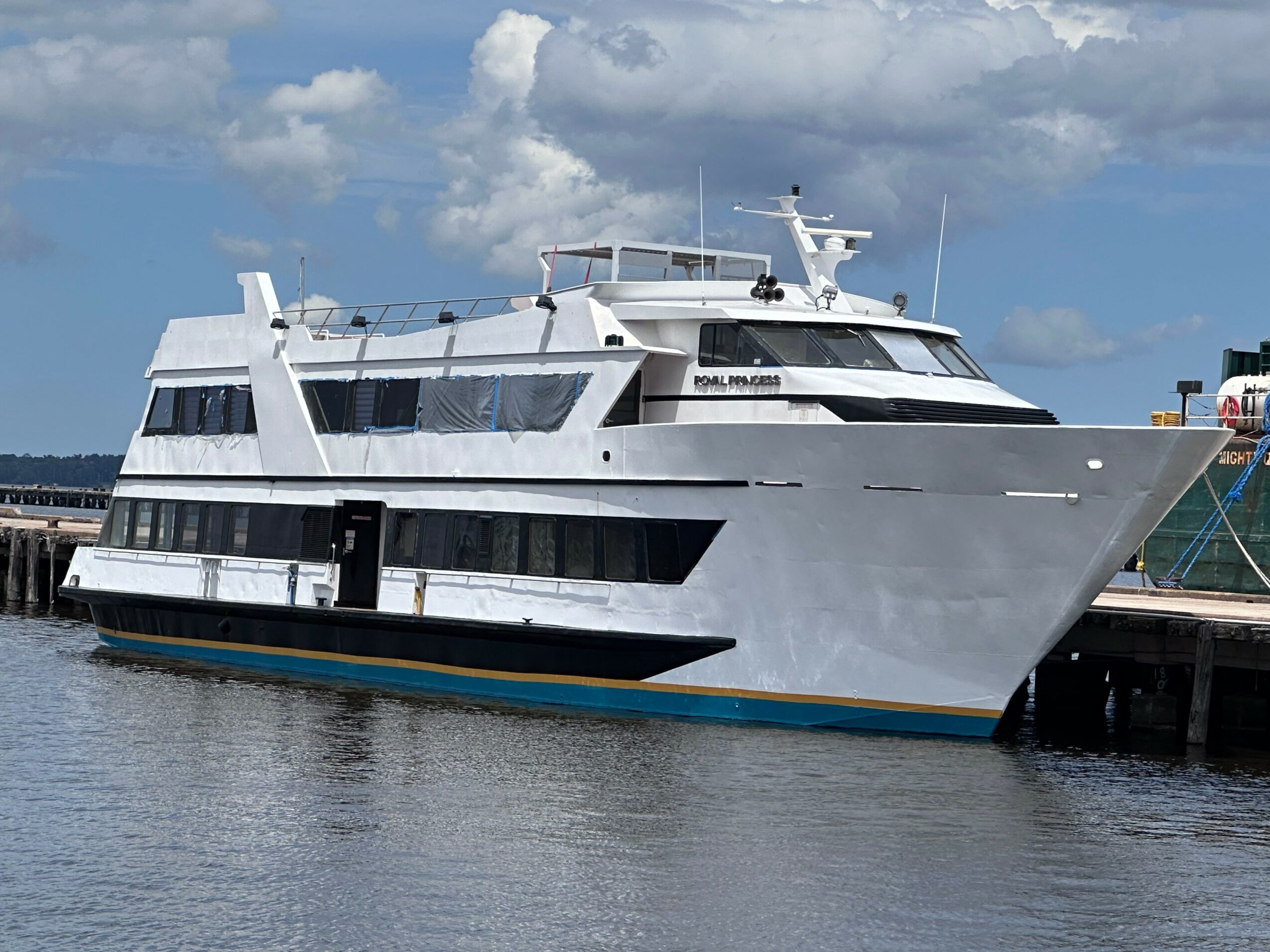 Large white yacht docked at a pier with water and cloudy sky in background.