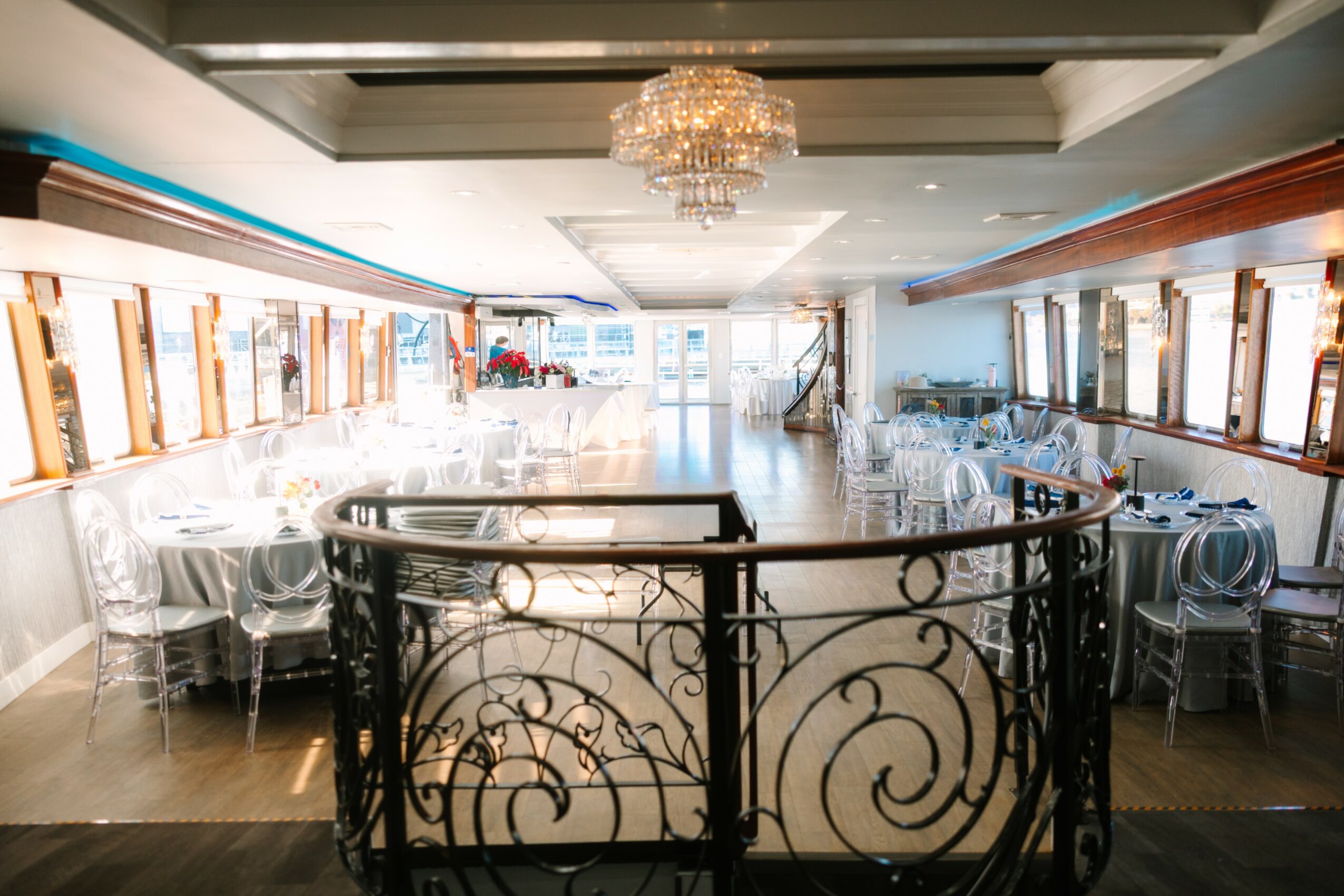 Elegant dining room with a chandelier, large windows, and tables set with glassware and chairs, viewed from a balcony with decorative railing.