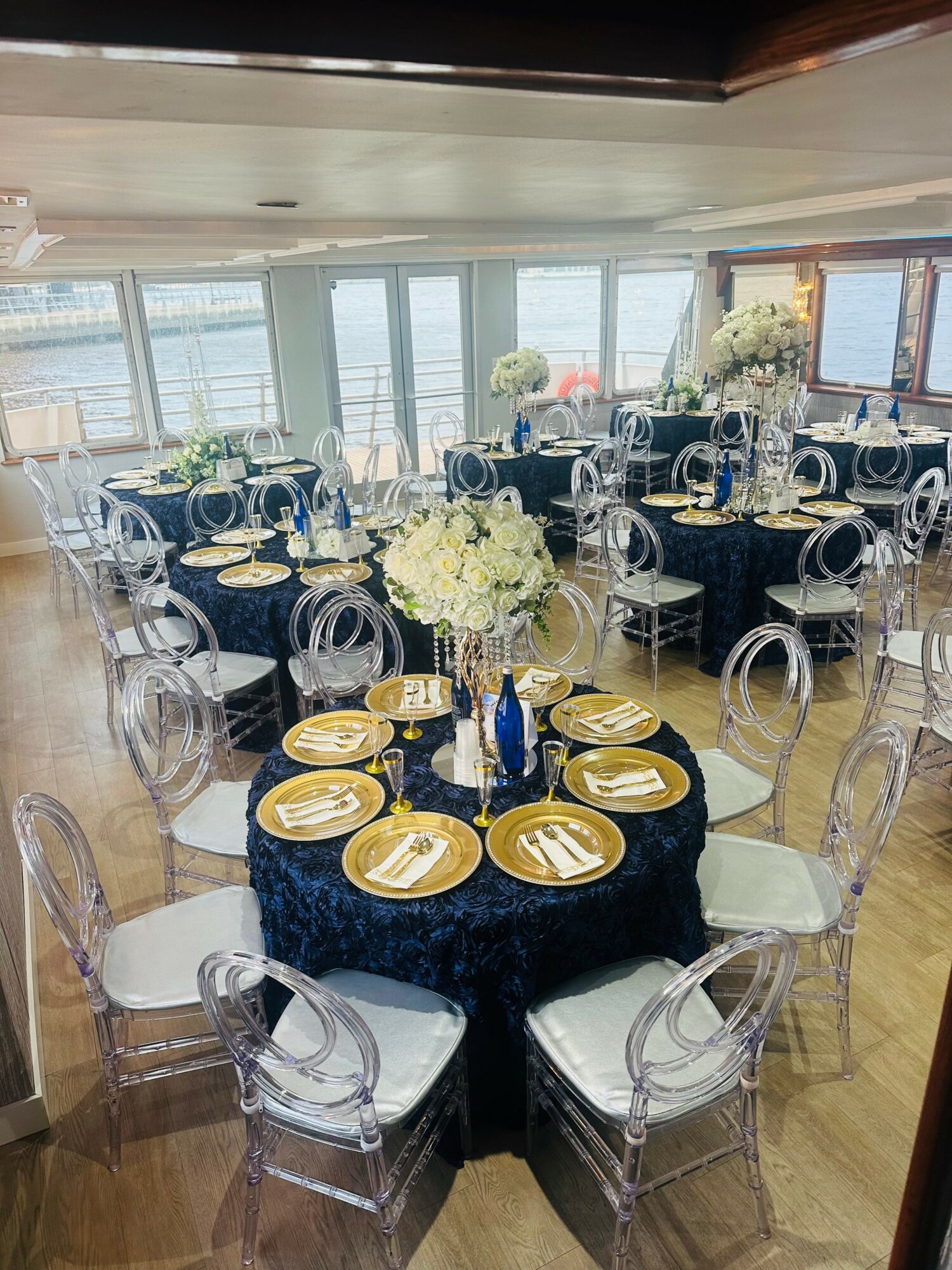 Elegant dining area with round tables, black tablecloths, white chairs, and floral centerpieces, overlooking a waterfront view.