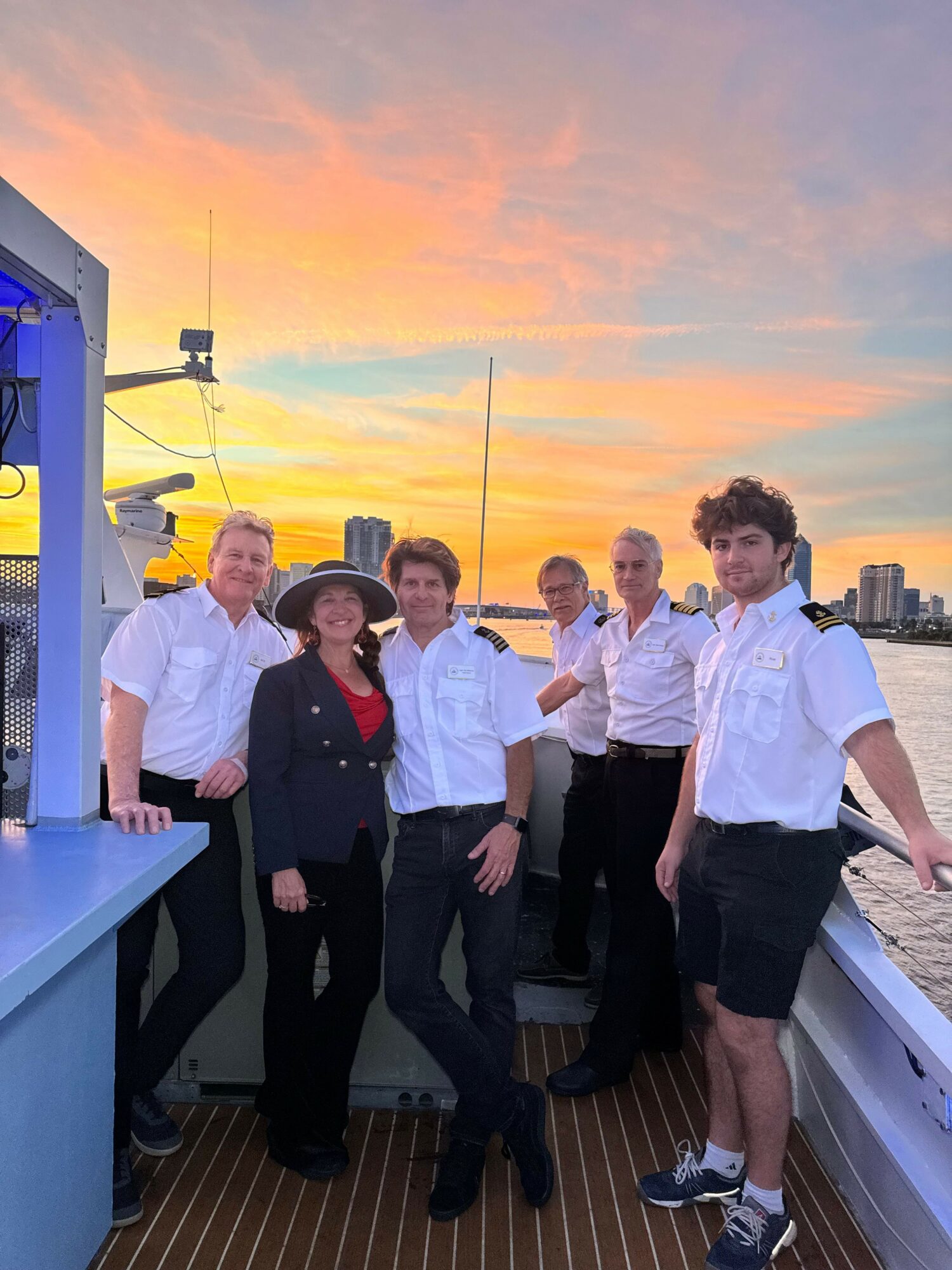 Group of people standing on a boat deck during sunset, with a city skyline in the background.