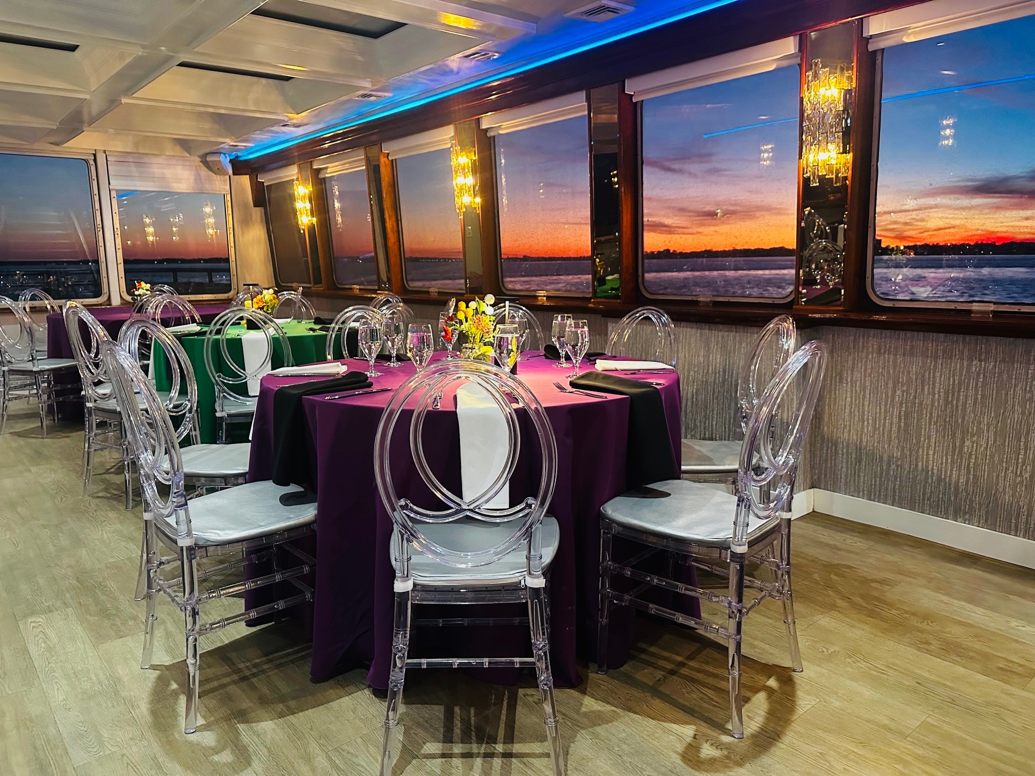Elegant dining area with round tables, purple tablecloths, clear chairs, and large windows showing a sunset view.