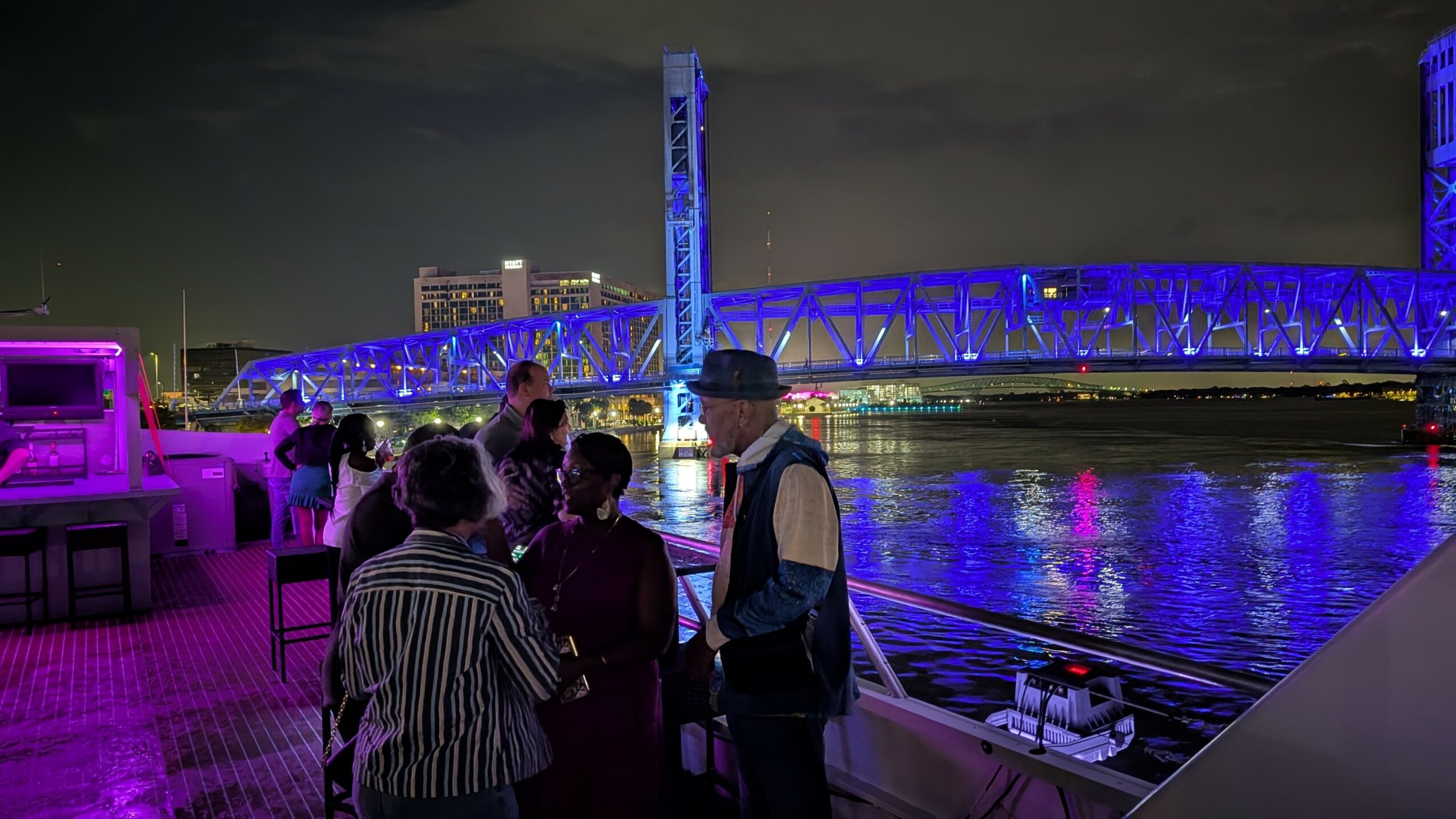 Group of people on a boat at night with a lit bridge and city skyline in the background.