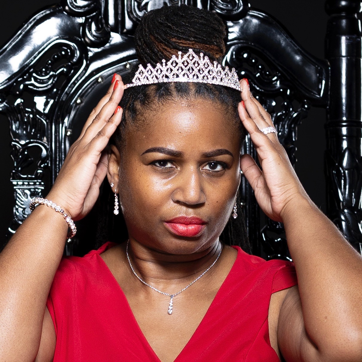 Woman wearing a tiara and red dress, holding her head, sitting on a black ornate chair, with jewelry and makeup.