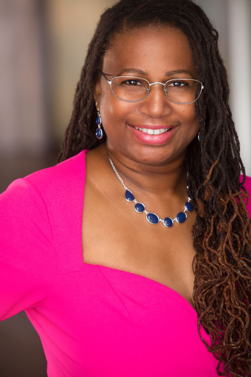 Woman with glasses and long dreadlocks wearing a pink dress and blue jewelry, smiling.