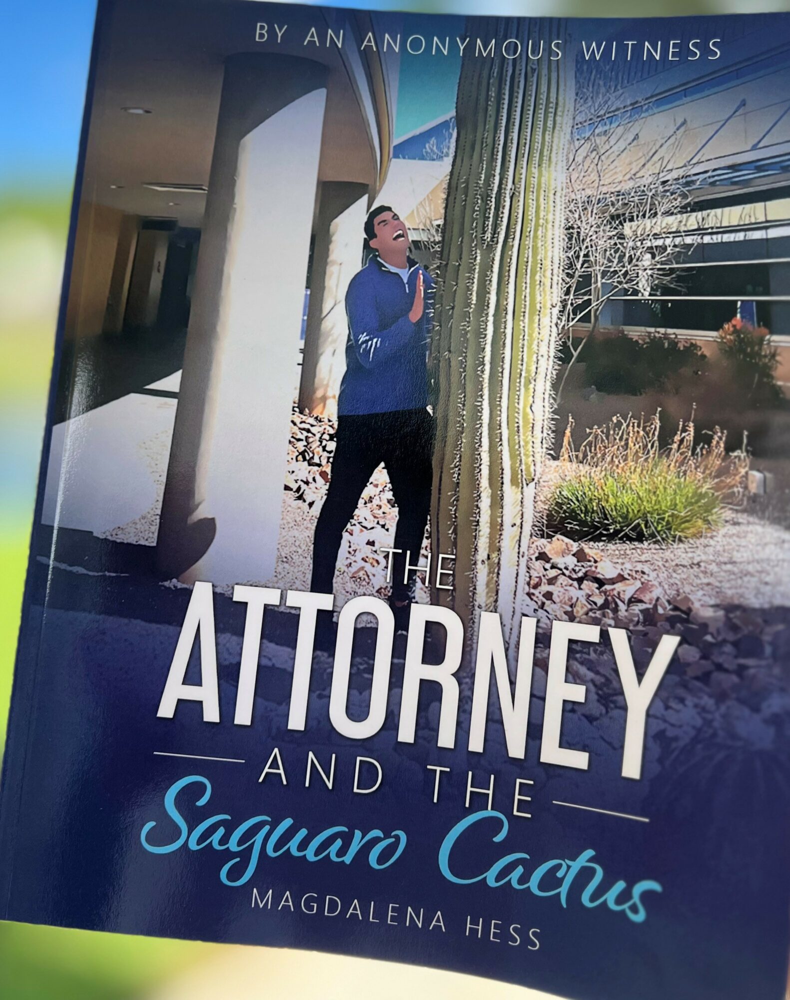 Person standing next to a large cactus outdoors, with a building and desert landscape in the background.