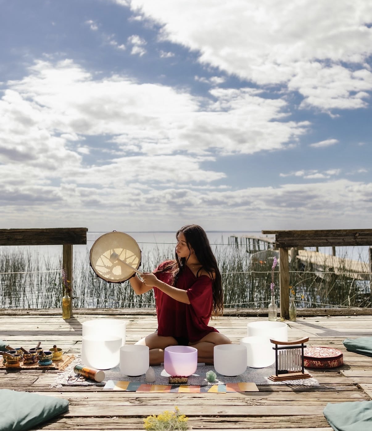 Girl sitting on wooden deck with bowls and decorations, holding a round object, outdoors under cloudy sky.
