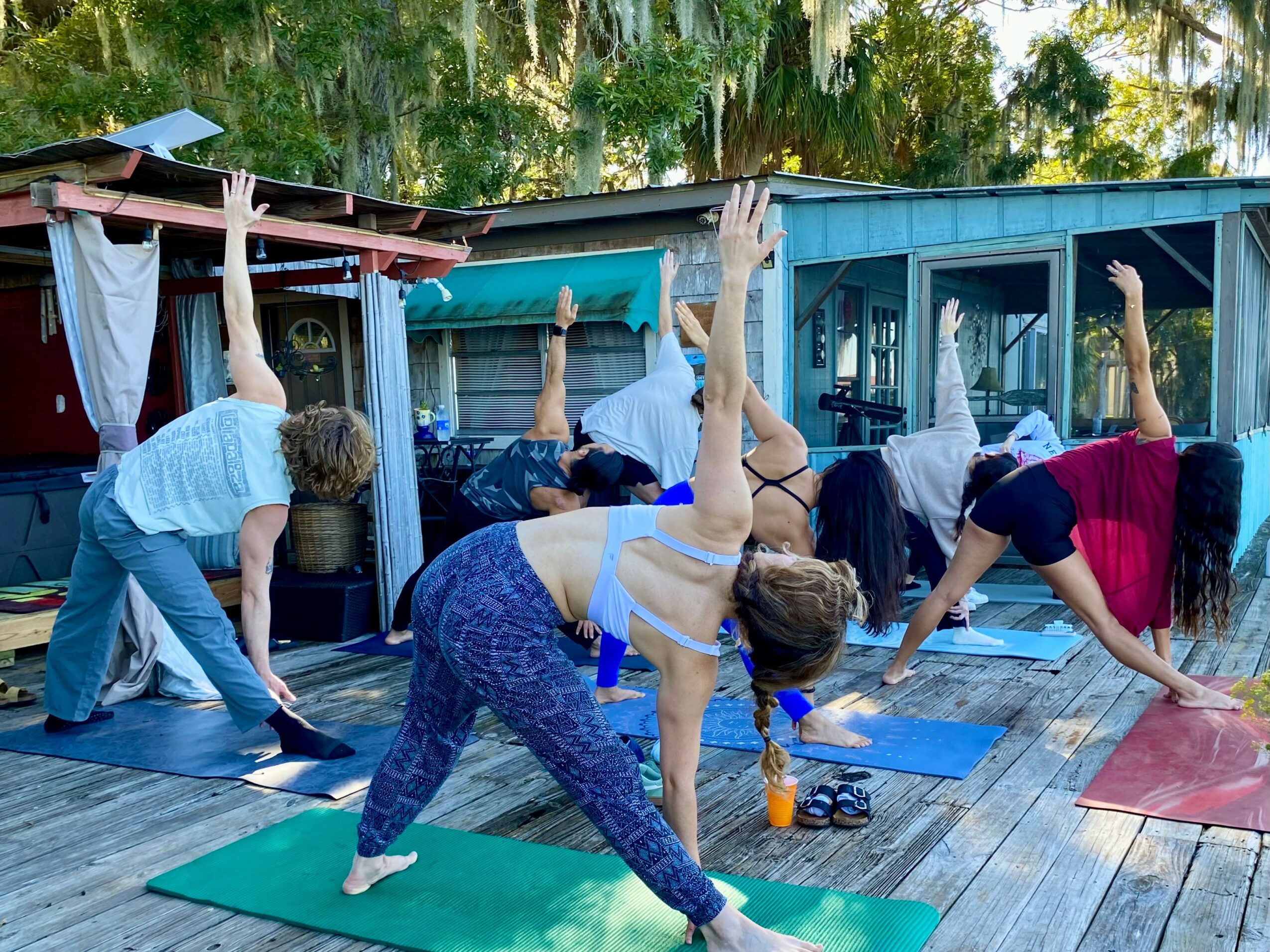 Group of people practicing yoga outdoors on mats, with trees and a building in the background.