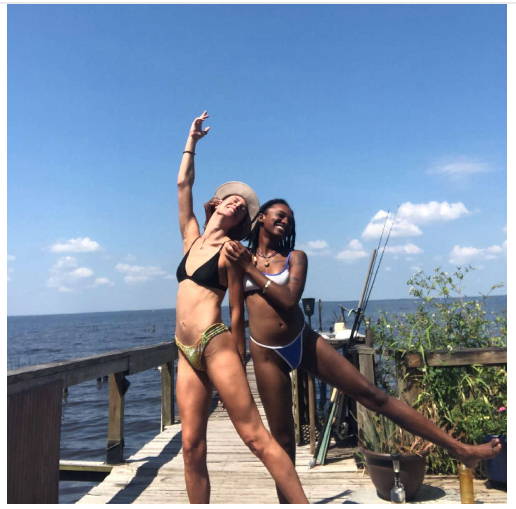 Two women in swimsuits pose on a wooden dock by the water, one raising an arm, the other extending a leg. Bright sky.