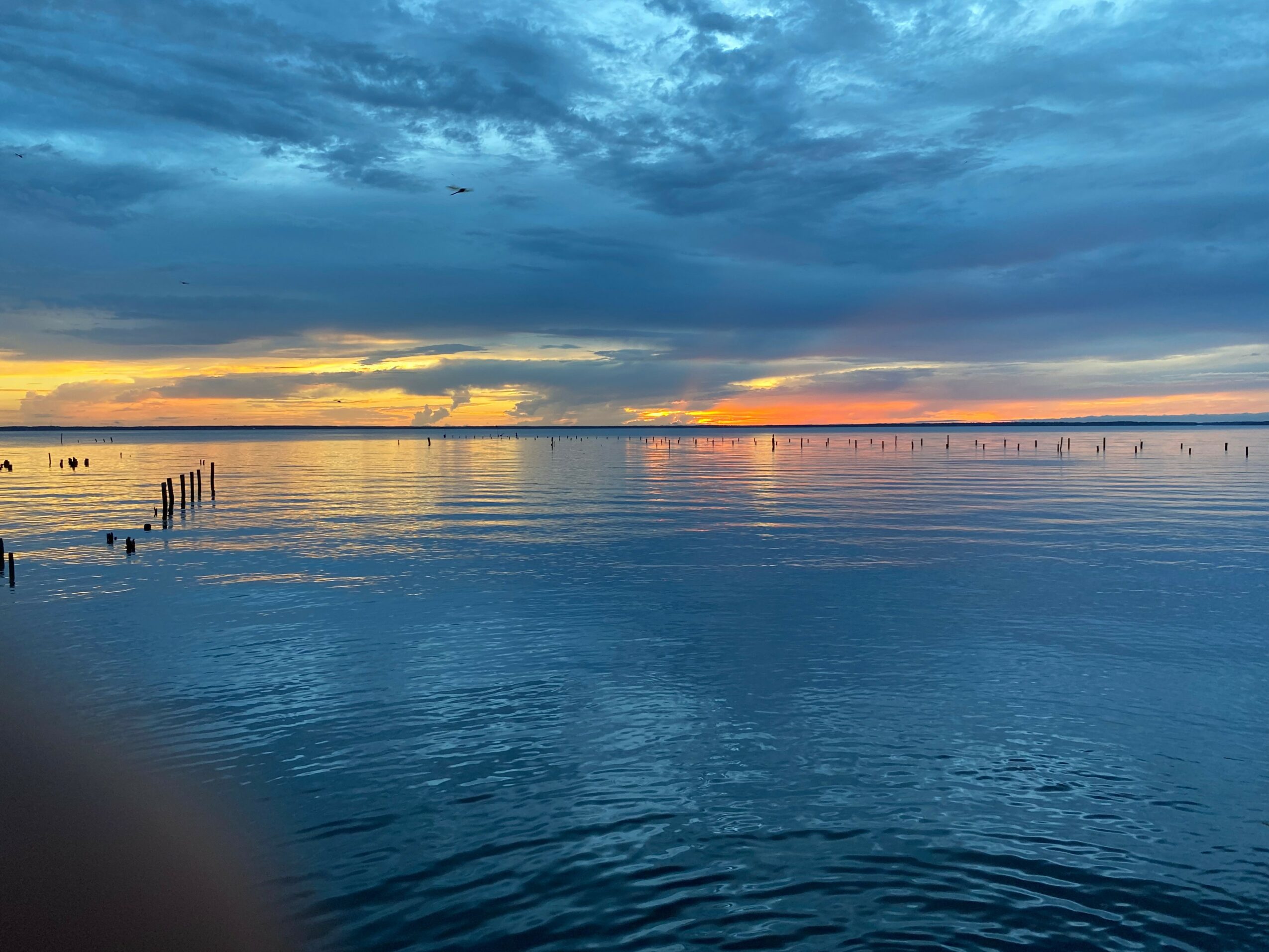 Sunset over calm water with a partly cloudy sky, orange and yellow hues near the horizon, and wooden posts in the water.