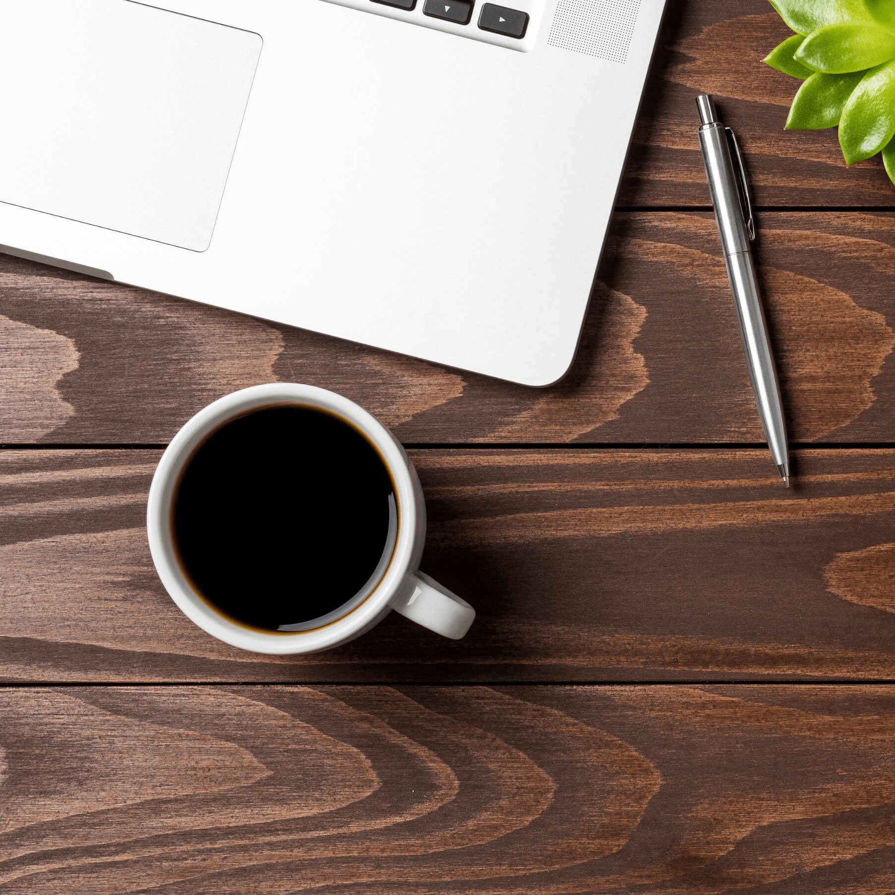 Top view of a coffee cup, laptop, pen, and plant on a wooden surface.