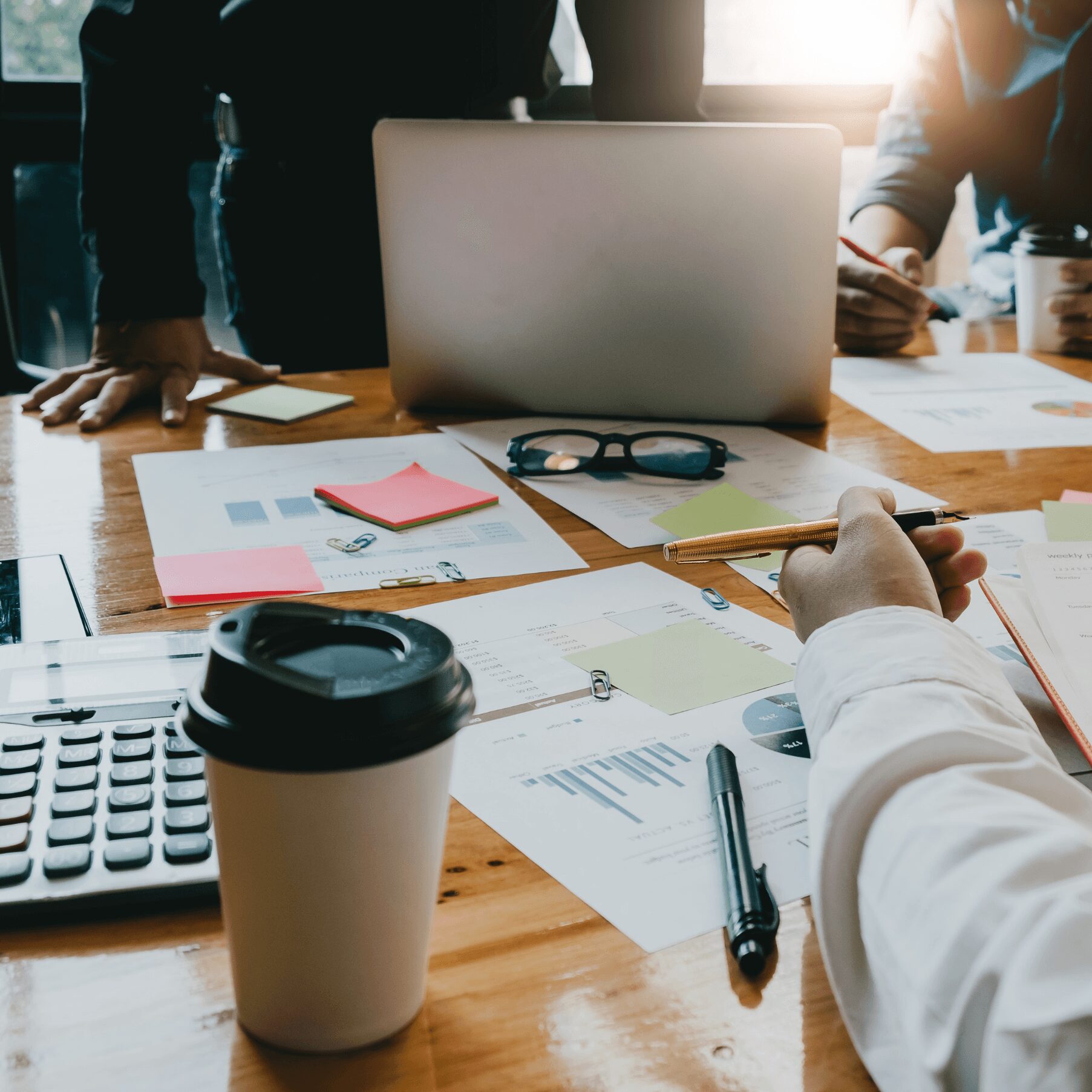 People working at a table with documents, a laptop, and a coffee cup, in a meeting or collaborative setting.