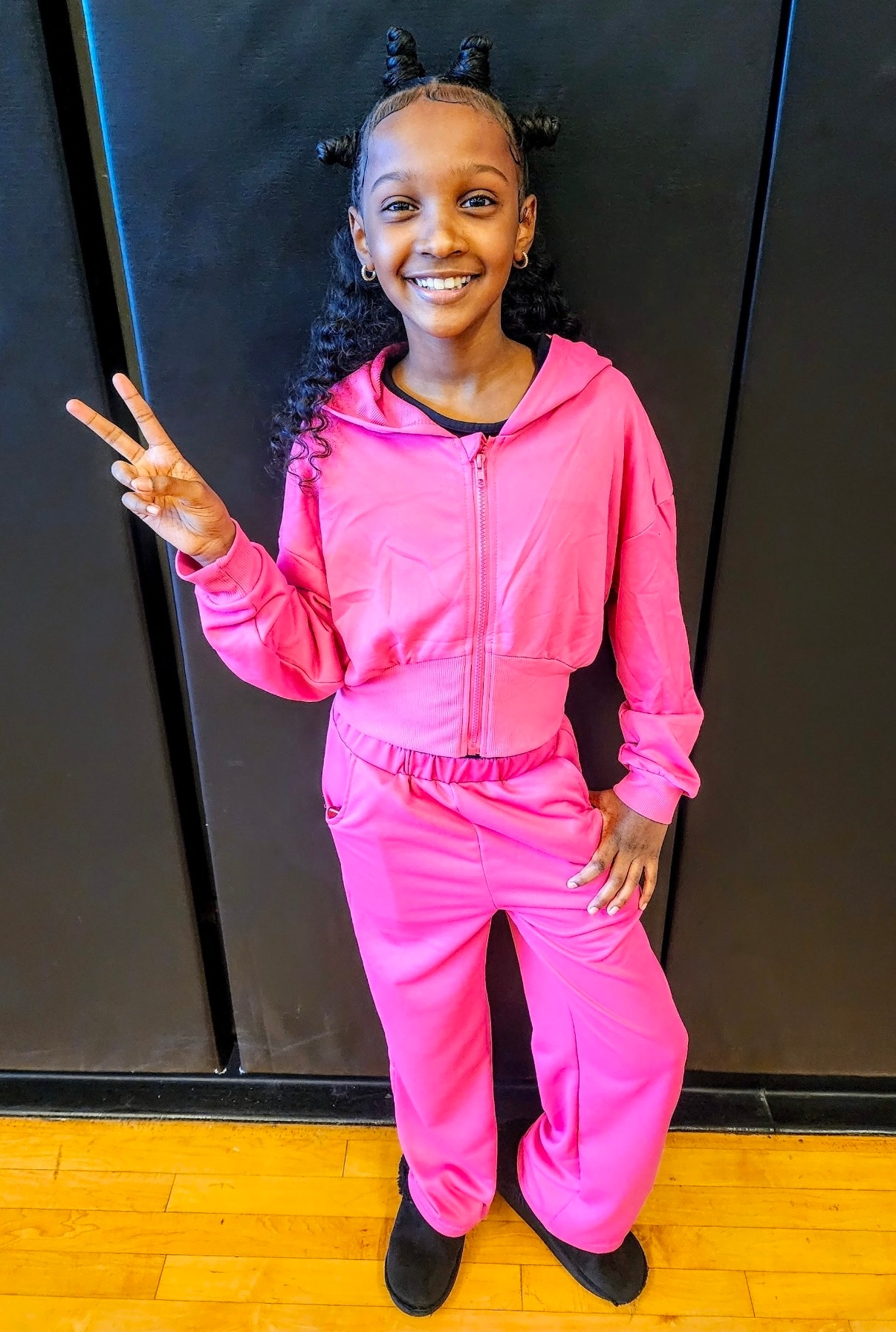 Smiling girl in pink tracksuit making peace sign, standing against black background, with wooden floor visible.