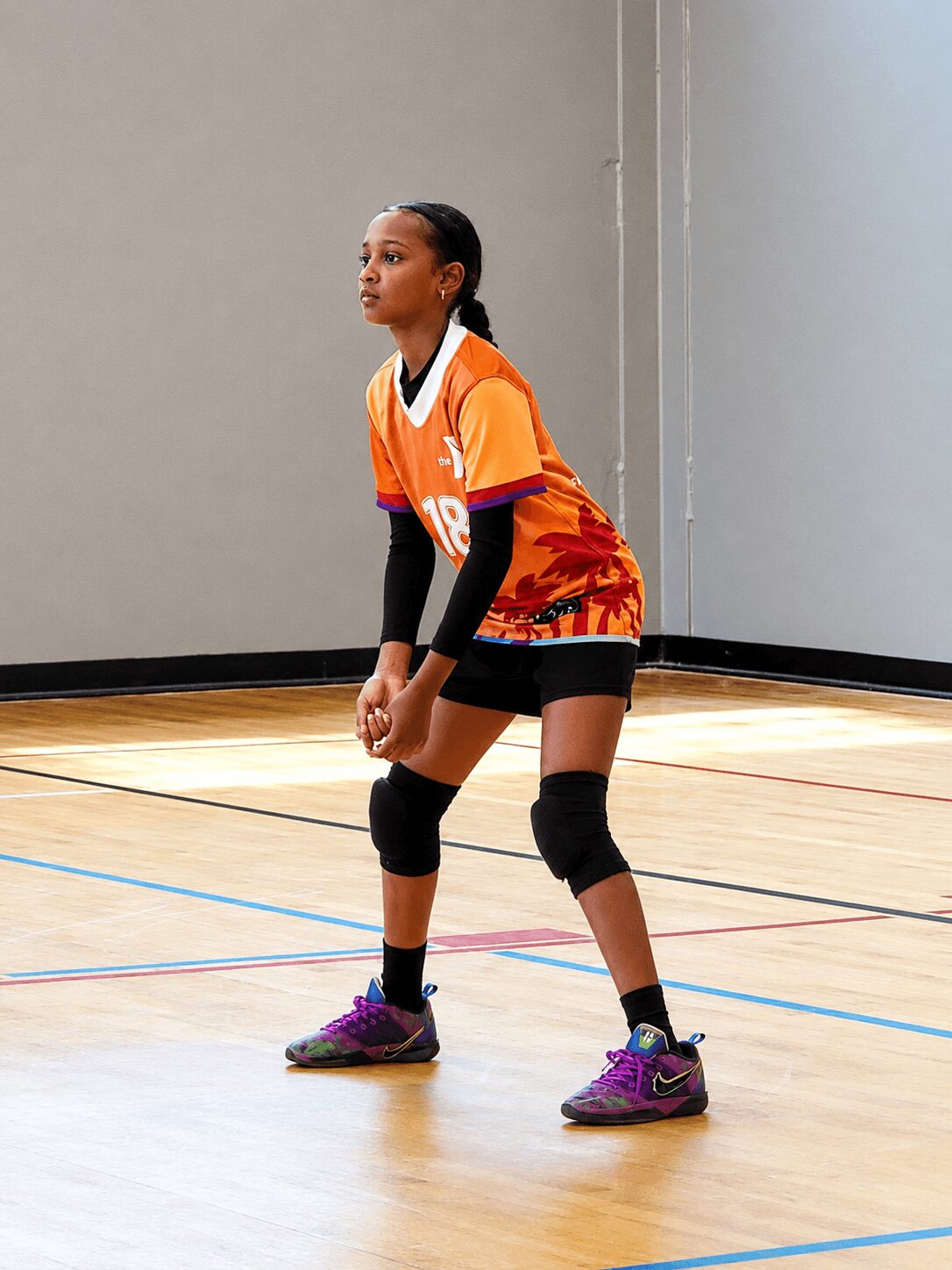 Young female volleyball player in orange uniform with black knee pads, standing on indoor court, ready to serve or receive.