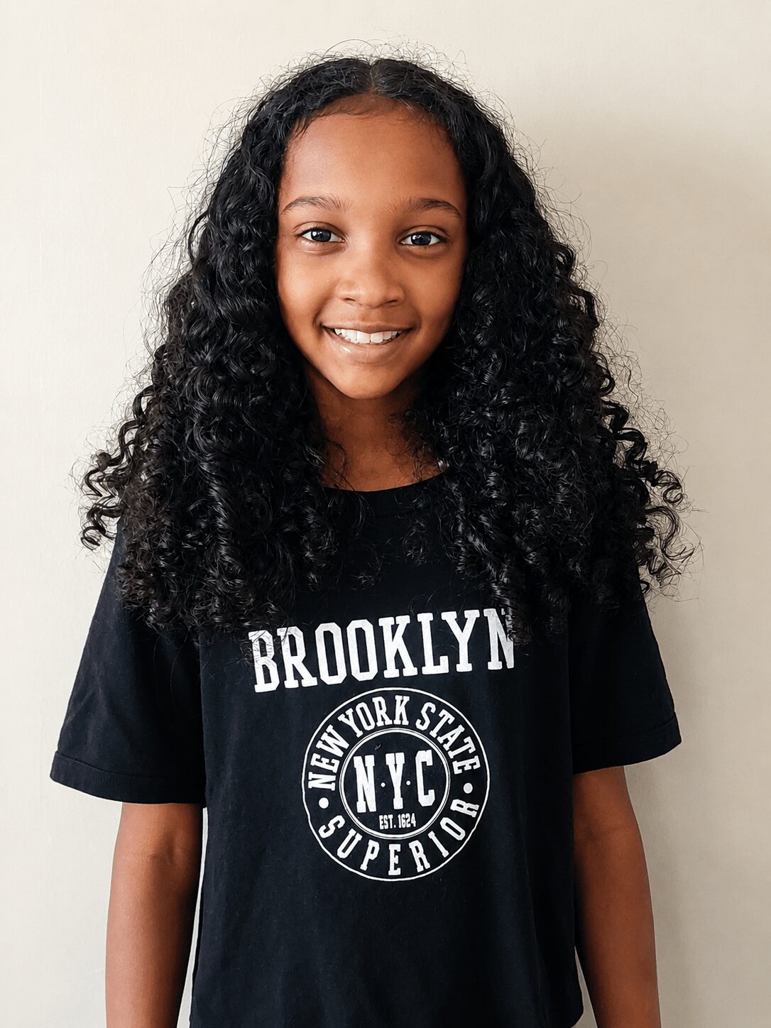 Smiling girl with curly hair wearing a black Brooklyn T-shirt, standing against a plain background.