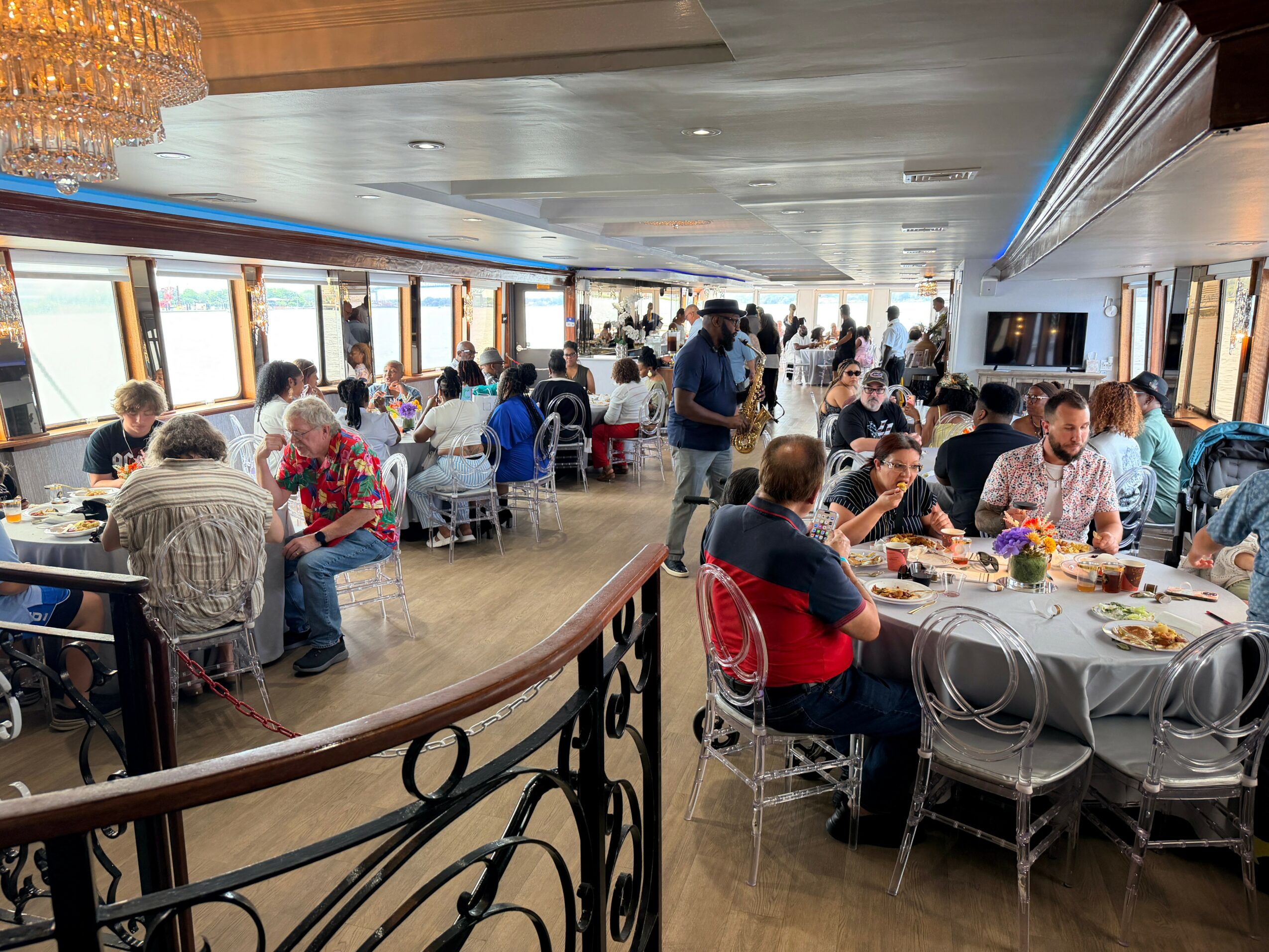 People dining in a large restaurant with windows, tables, and chairs, some standing and talking, others eating, with a chandelier visible.