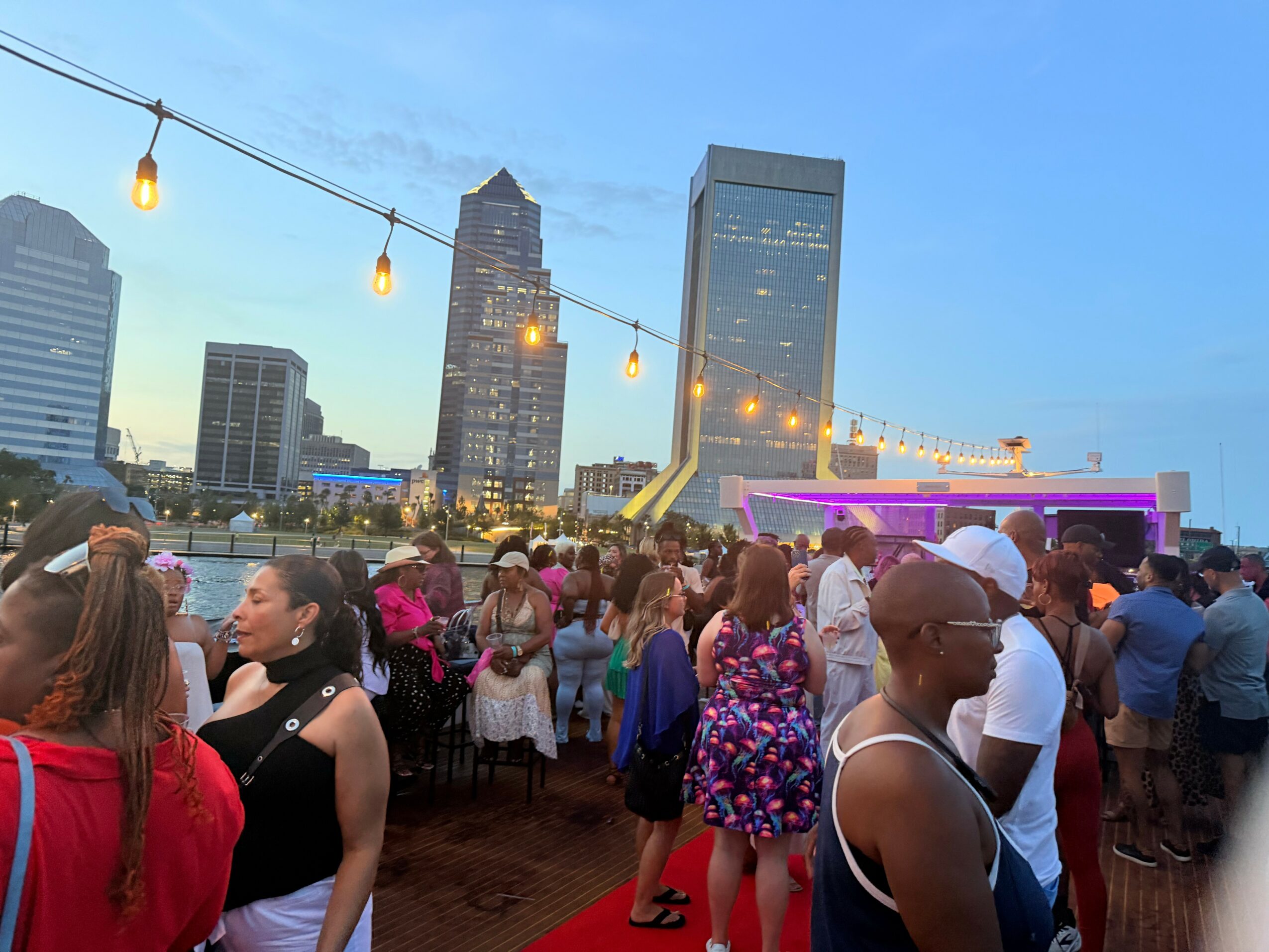 Crowd of people outdoors with city skyline and string lights in the background during evening.