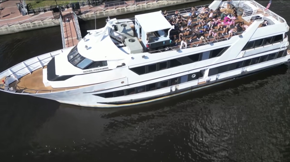Large white yacht on water with people on upper deck, surrounded by a cityscape and greenery.