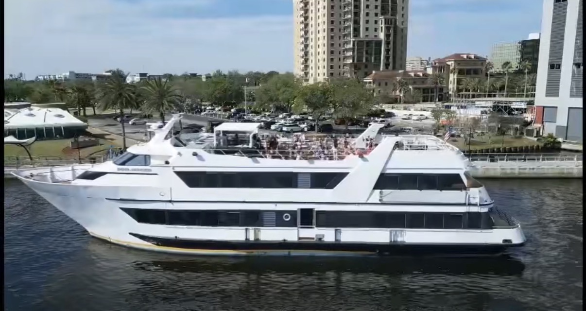 Large white yacht docked in water with buildings and trees in background.