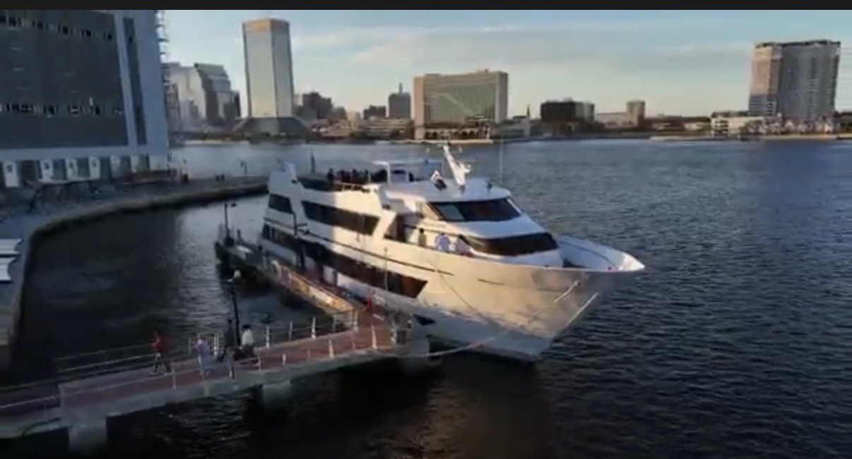 Large white yacht docked at a pier with city skyline in background, water reflecting sunlight.
