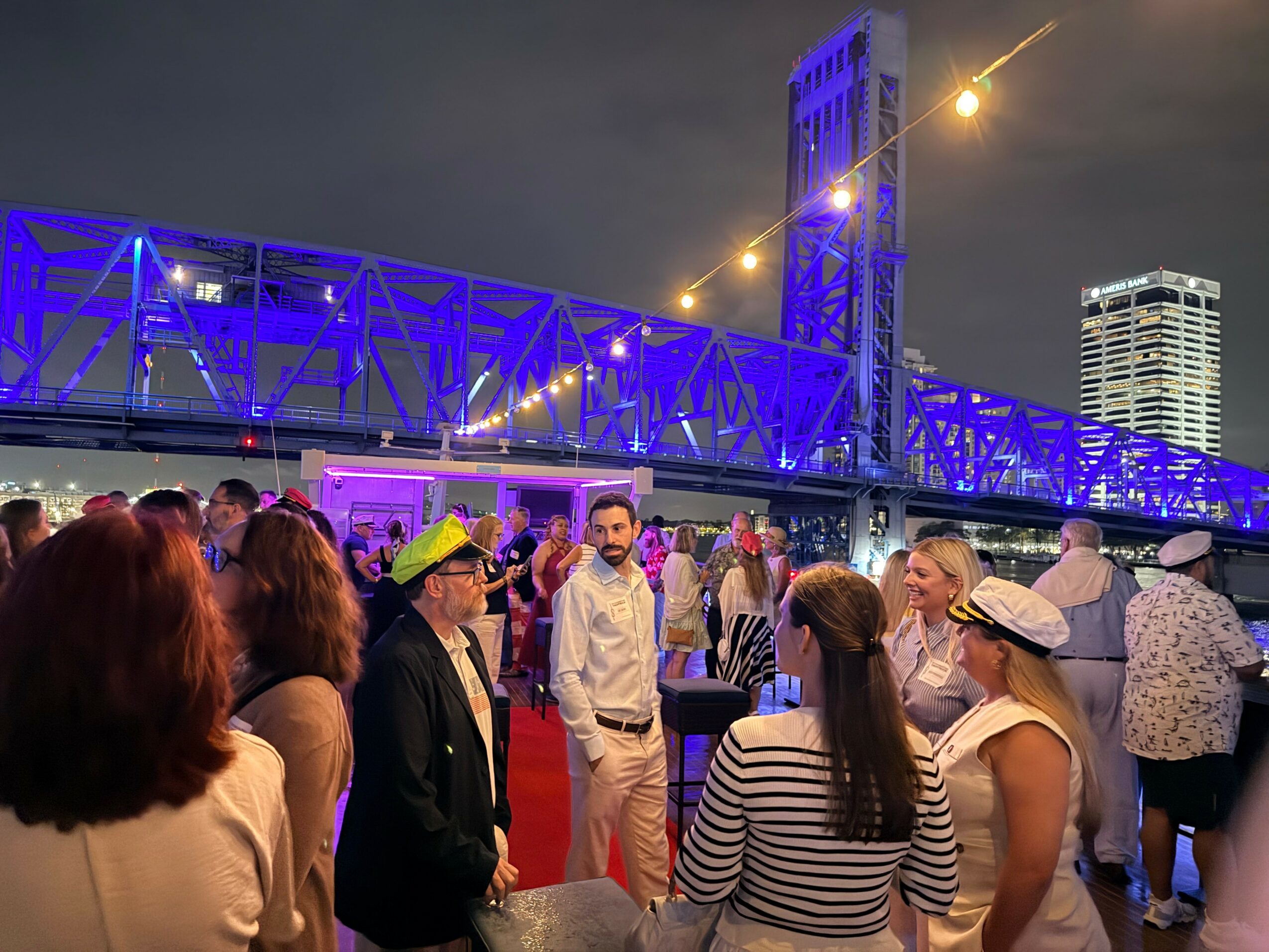 People gather outdoors at night near a bridge illuminated in blue, with city buildings in the background, some wearing hats and casual clothing.