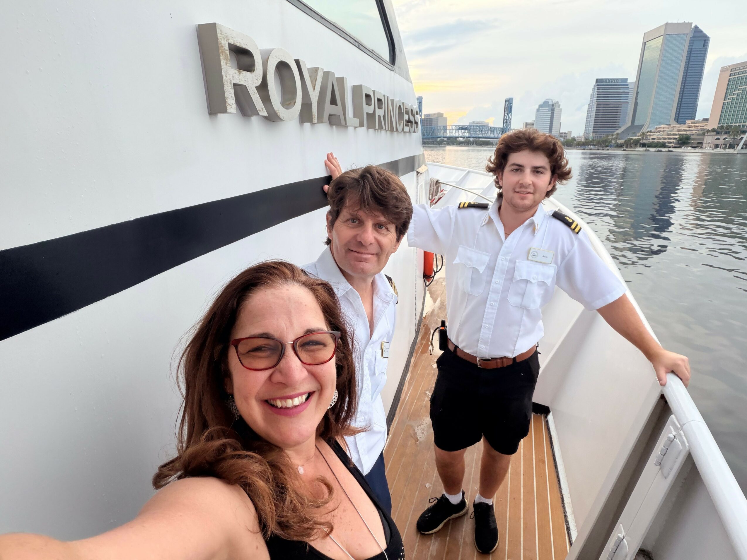 Three people on a boat with a city skyline and water in the background, smiling and taking a selfie.