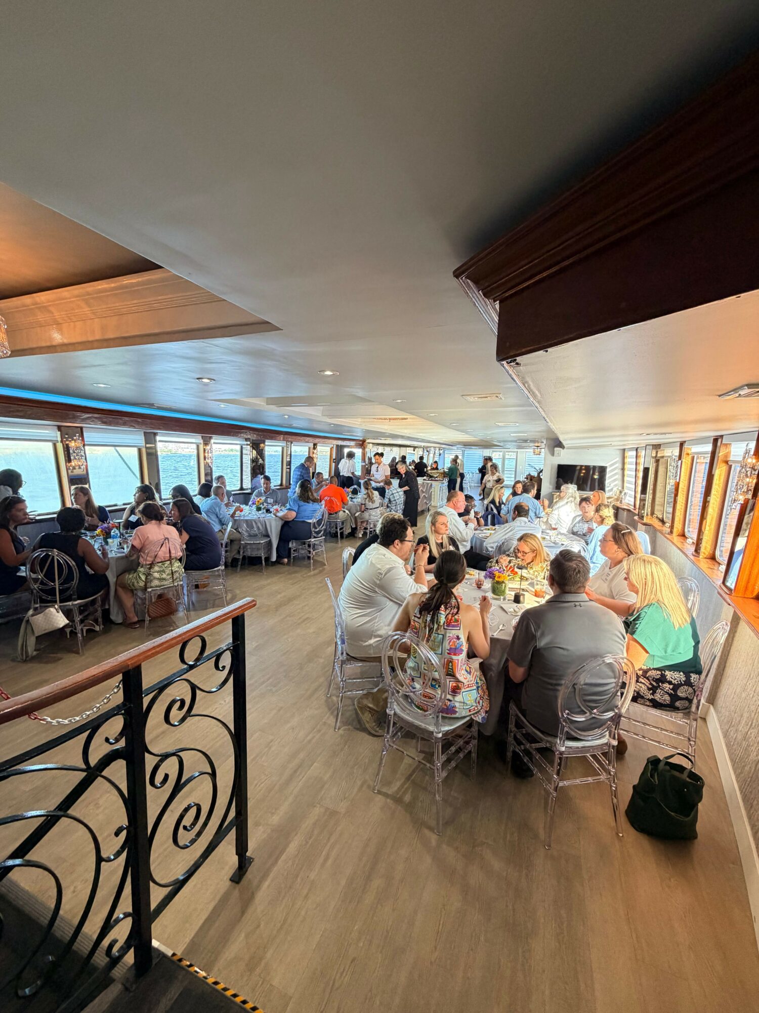 People gather in a room with large windows, sitting at tables and engaging in activities, with a wooden floor and decorative railing.