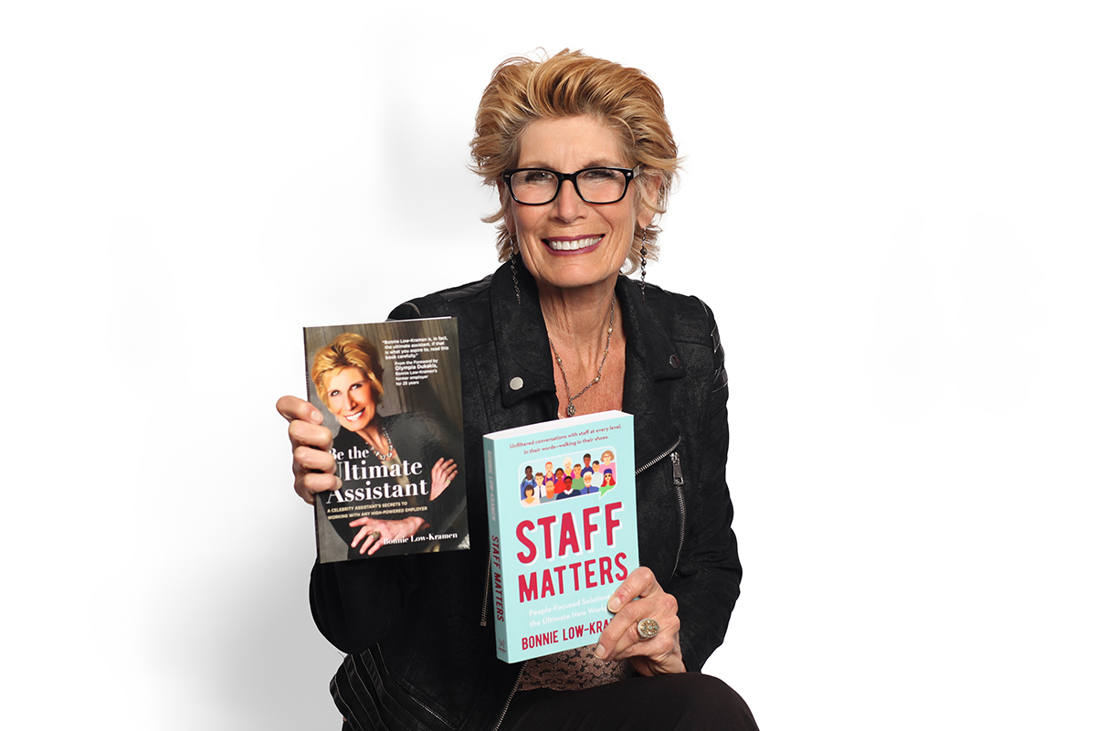 Smiling woman with glasses holding two books, one titled 'Staff Matters' and the other 'The Ultimate Assistant,' against a white background.