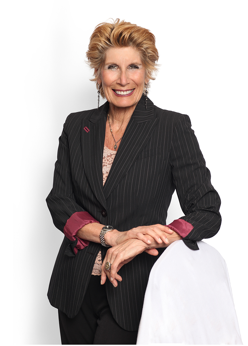 Smiling woman with short, wavy blonde hair wearing a black pinstripe blazer with pink cuffs, sitting at a table.