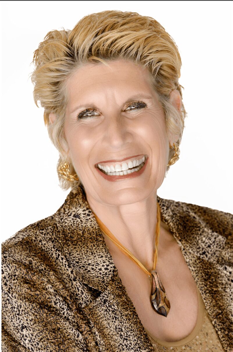 Smiling woman with short blonde hair, wearing a patterned jacket and gold jewelry, against a white background.