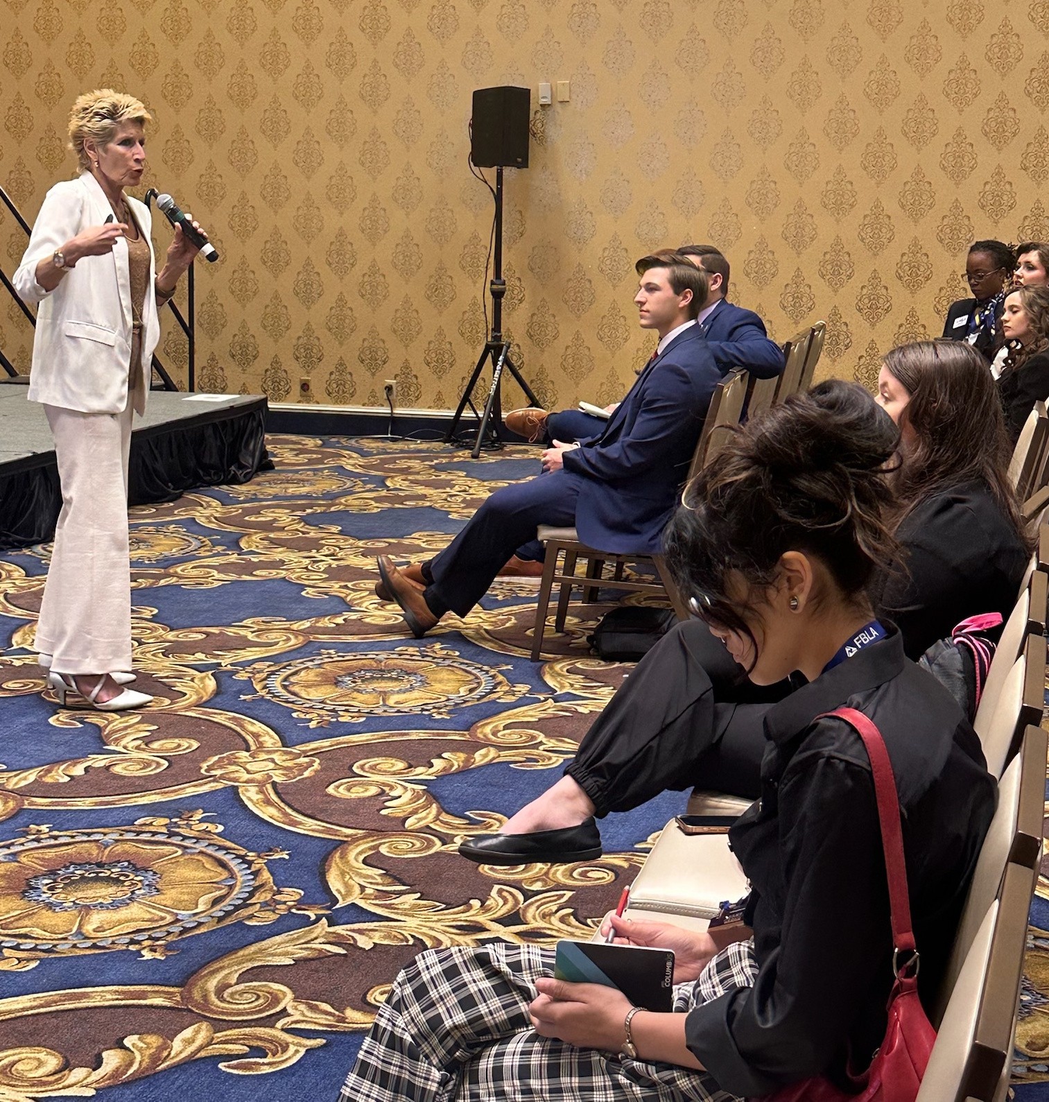 Woman speaking into microphone in front of seated audience in a conference room with patterned carpet and wallpaper.