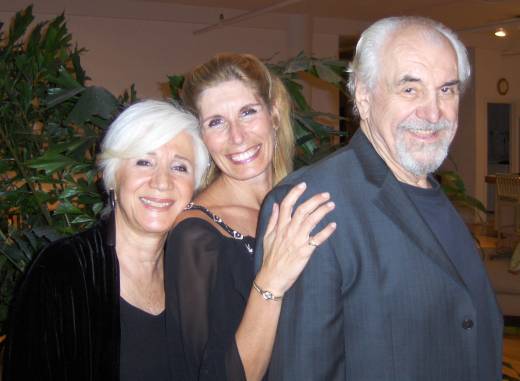 Three smiling people, two women and one man, standing close together indoors with plants in the background.