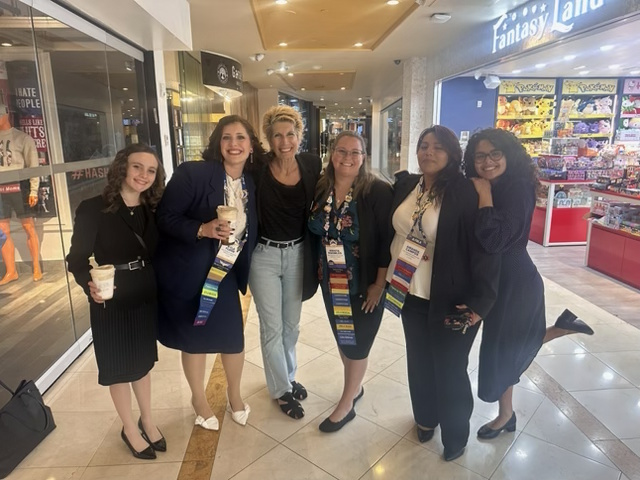Six women standing together in a shopping mall, smiling, holding drinks, and wearing colorful scarves, with stores in the background.