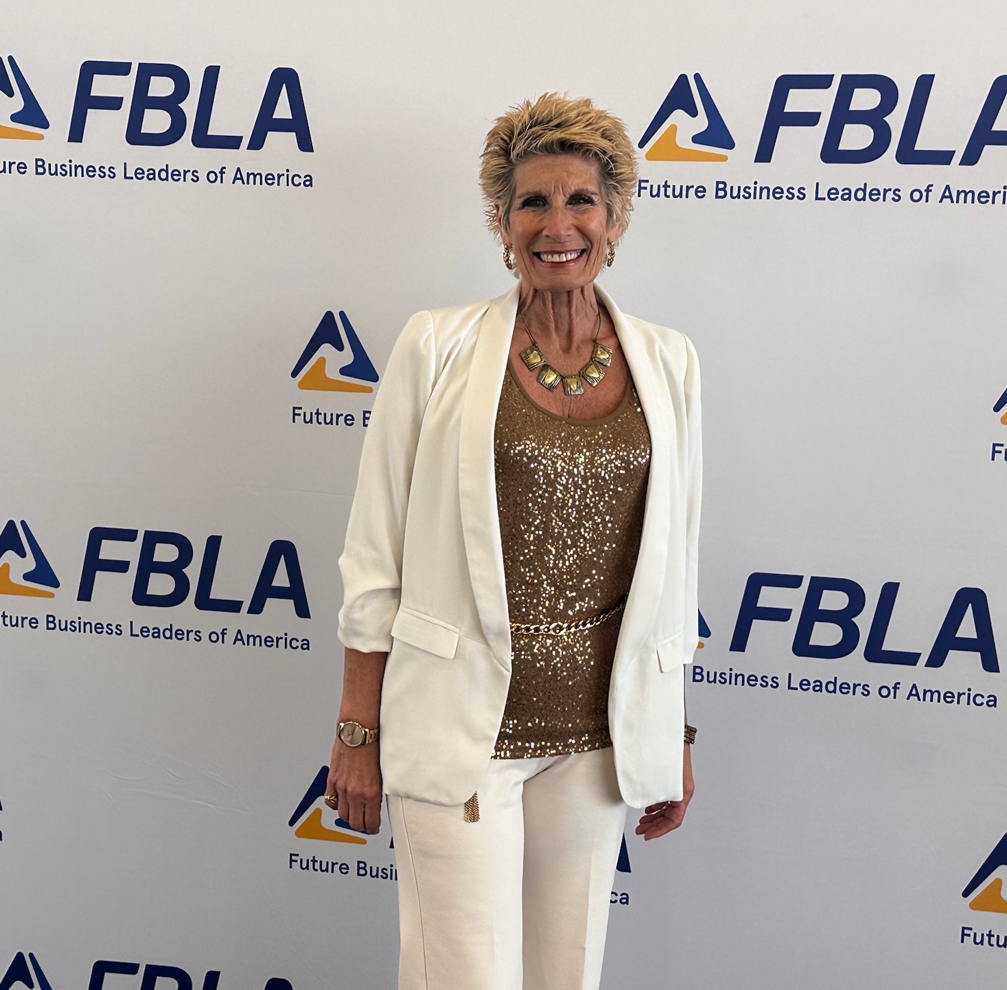 Smiling woman in white blazer and pants standing in front of FBLA backdrop, wearing a gold top and necklace.