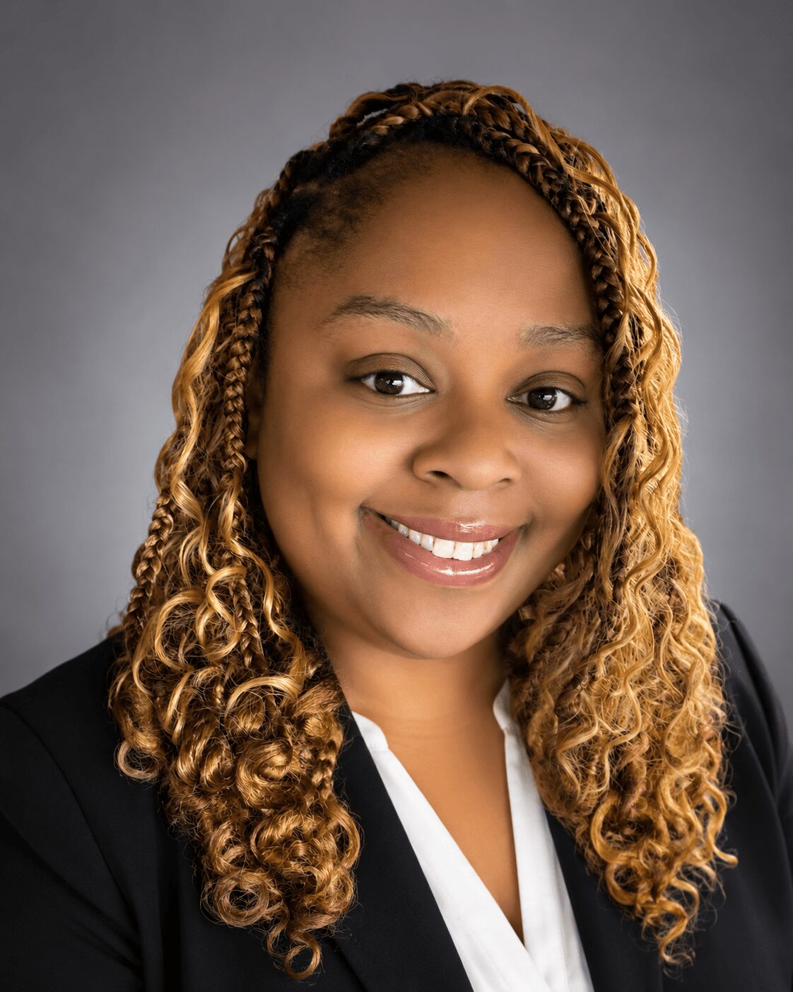 Portrait of a smiling woman with curly, blonde-tinted hair, wearing a black blazer and white top, against a gray background.