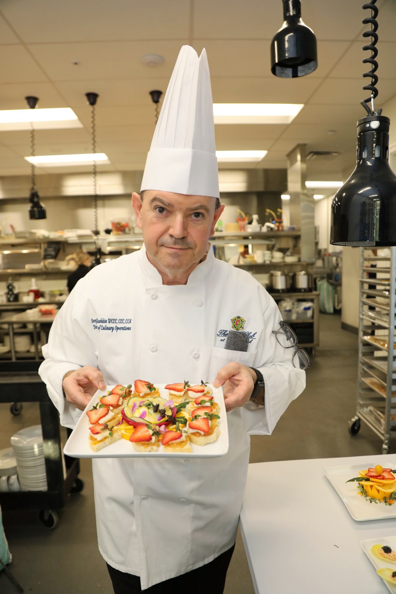 Chef in white uniform and tall hat holding a plate of colorful food in a professional kitchen.