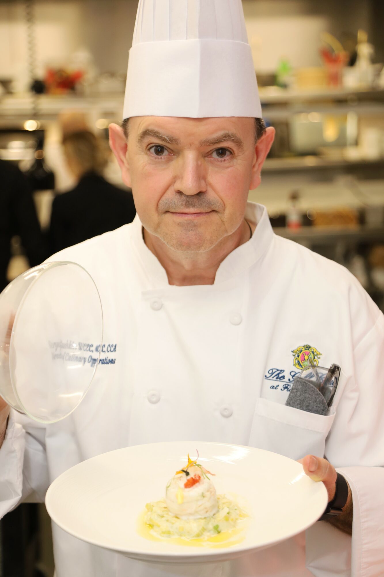 Chef in white uniform and hat holding a plate with a dish, kitchen background, focused expression.