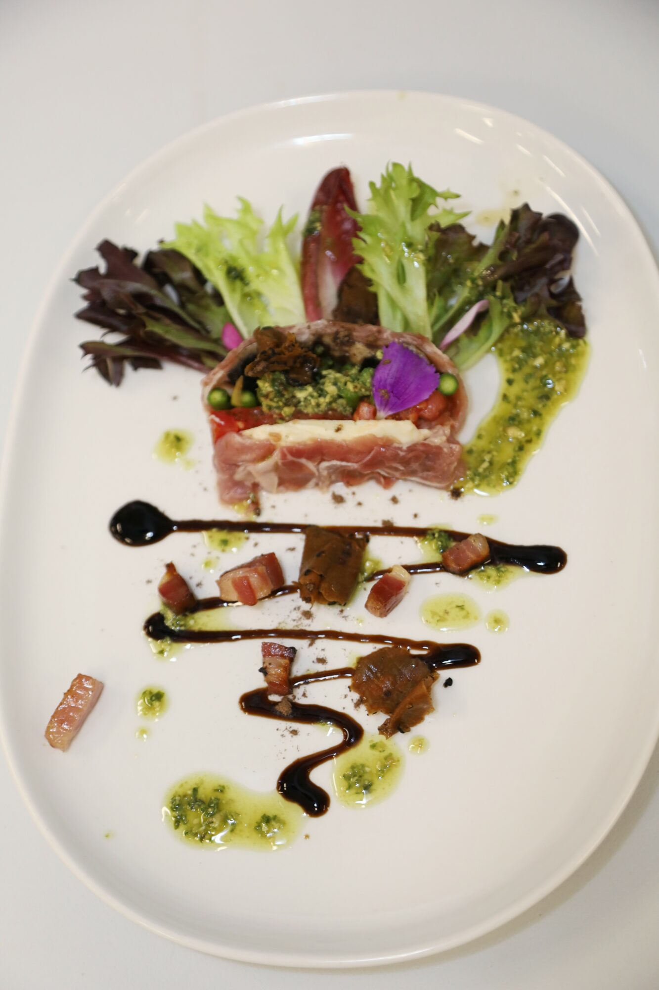 Plate with layered meat and vegetables, garnished with greens, purple flower, and drizzled sauces, on a white oval dish.