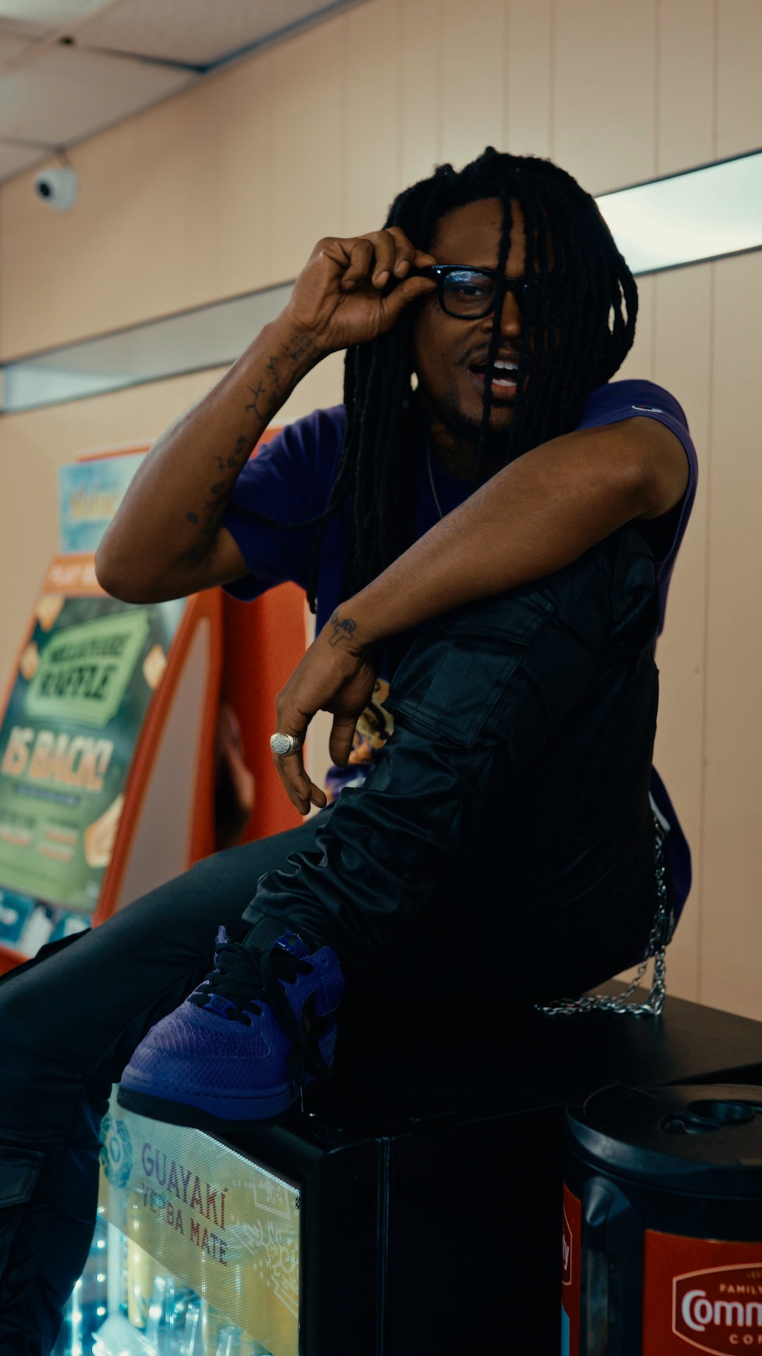 Person with glasses and dreadlocks adjusting glasses, sitting with crossed arms, in an indoor setting.
