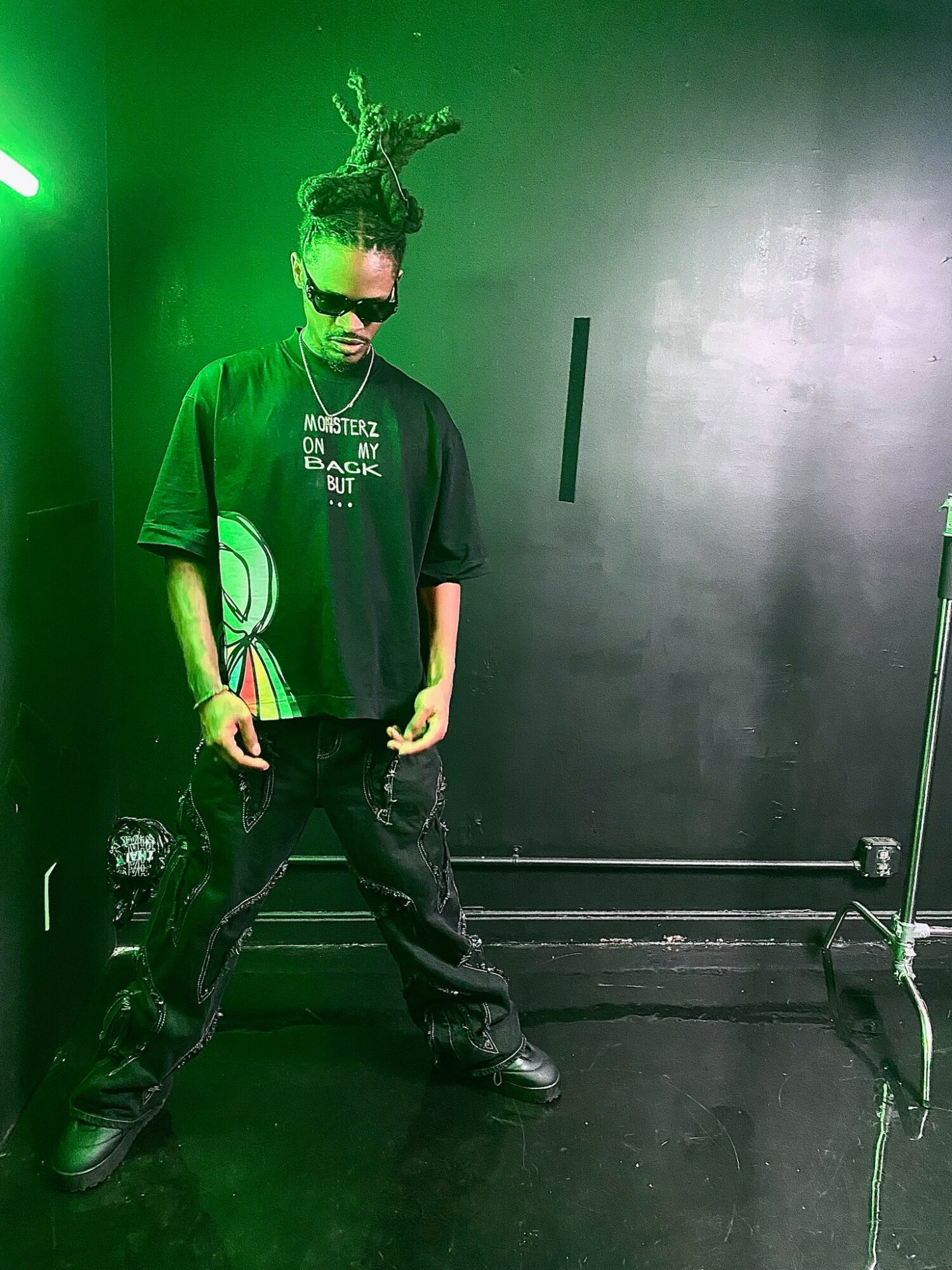 Person with dreadlocks, sunglasses, and black clothing stands in a dark room with green lighting, posing confidently.