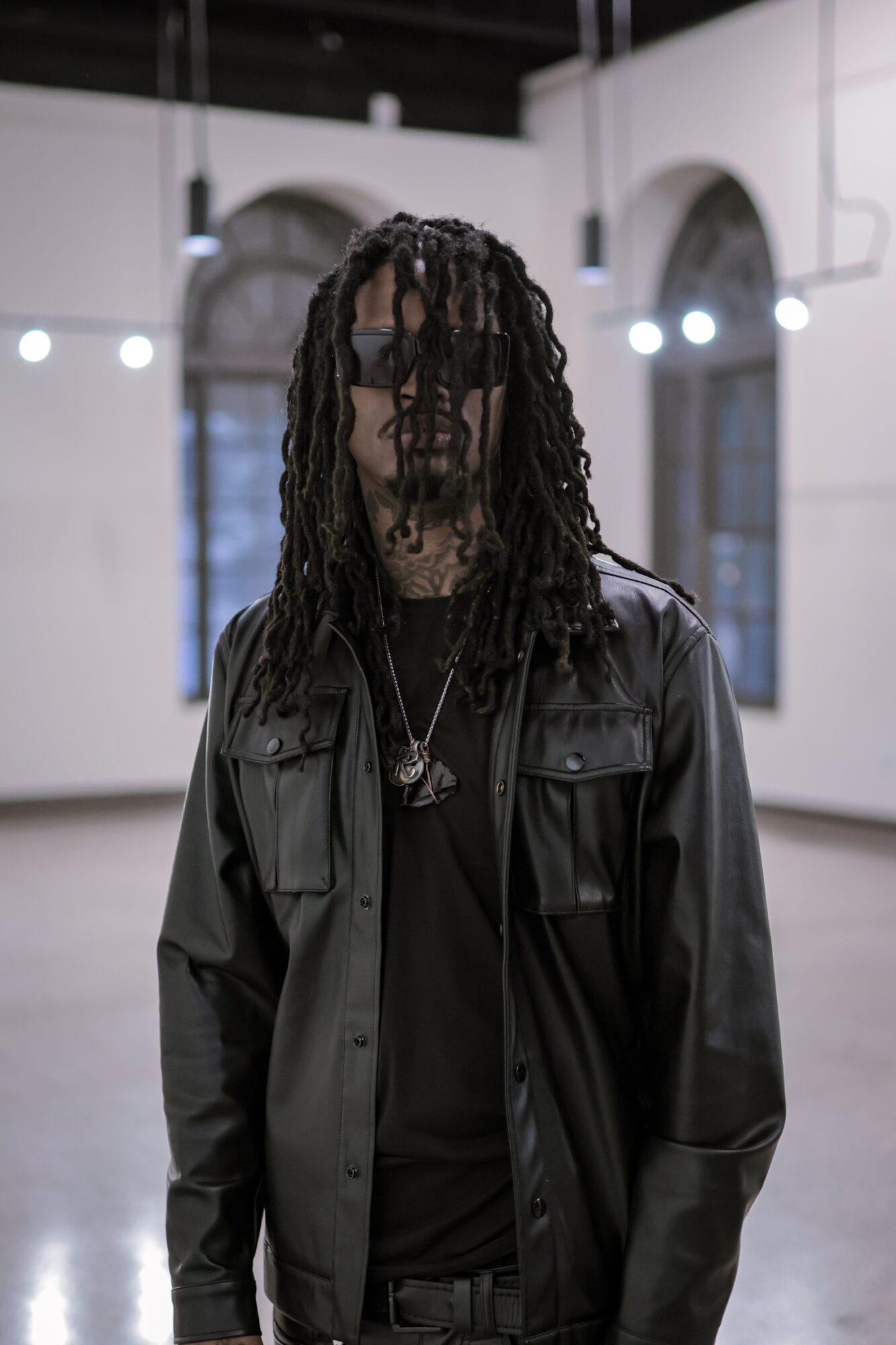 Person with long dreadlocks wearing a black leather jacket and sunglasses indoors, with bright lights and arched windows in background.