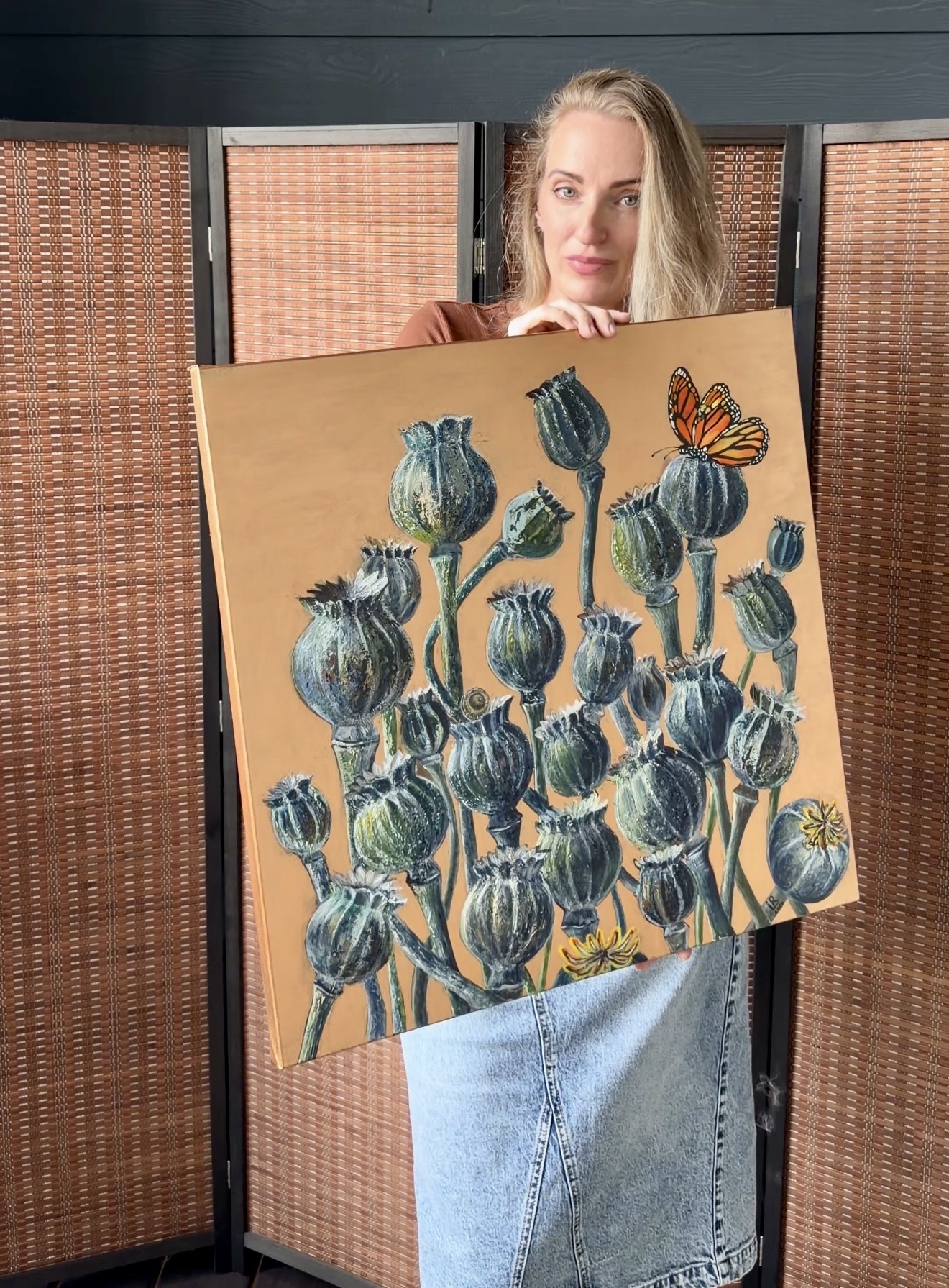 Woman holding a painting of blue flowers and a butterfly, sitting behind a folding screen.