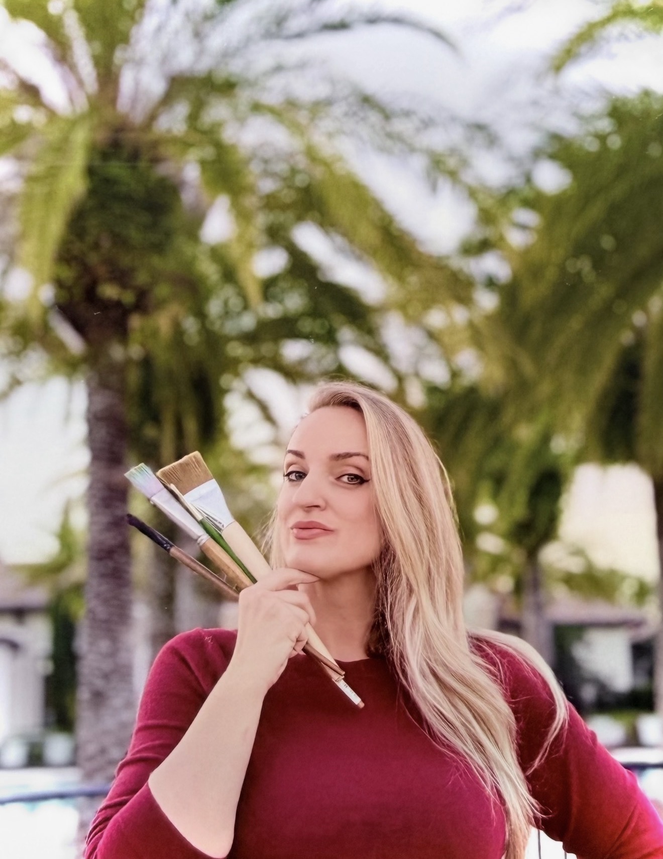 Woman with long blonde hair in a red top outdoors, holding paintbrushes, with palm trees in background.