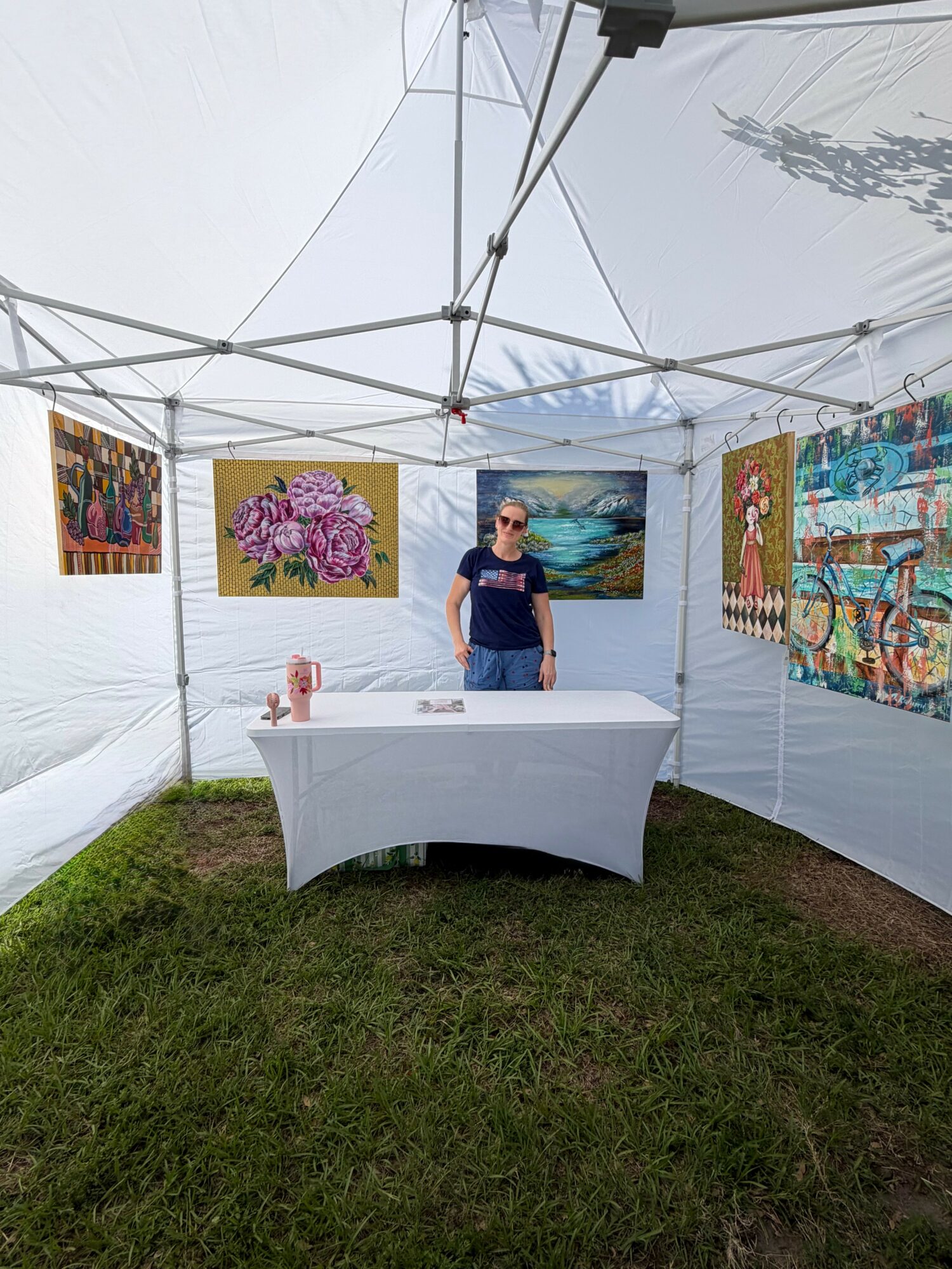 Person standing behind a table inside an art tent with colorful paintings on the walls.