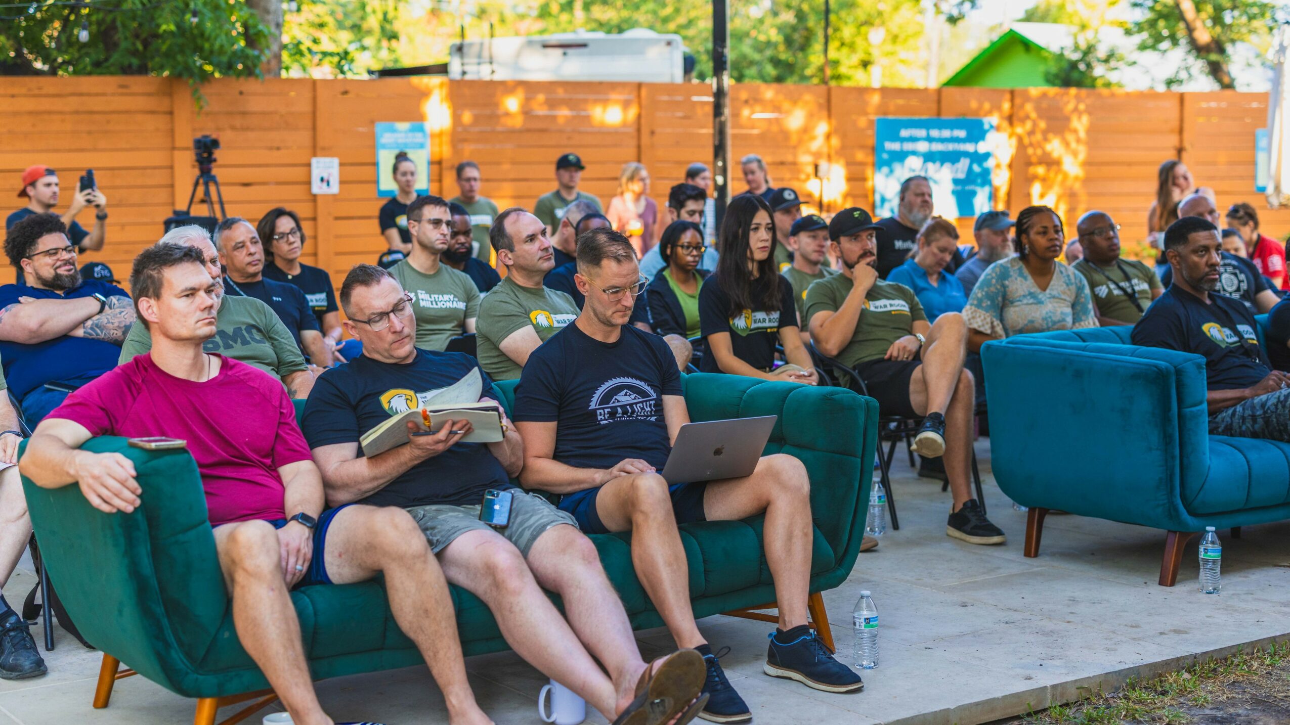 Group of people seated outdoors, some using laptops, with a wooden fence and trees in the background.