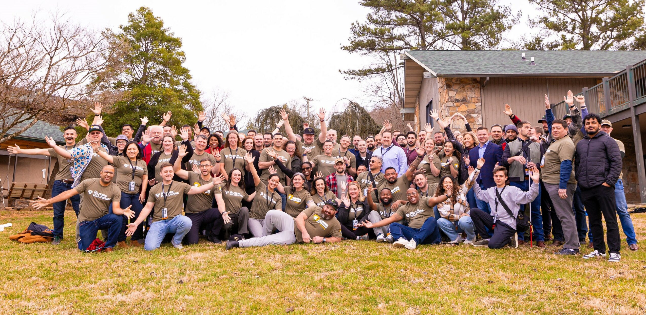 Large group of people outdoors in front of a house, some with arms raised, smiling, during daytime.