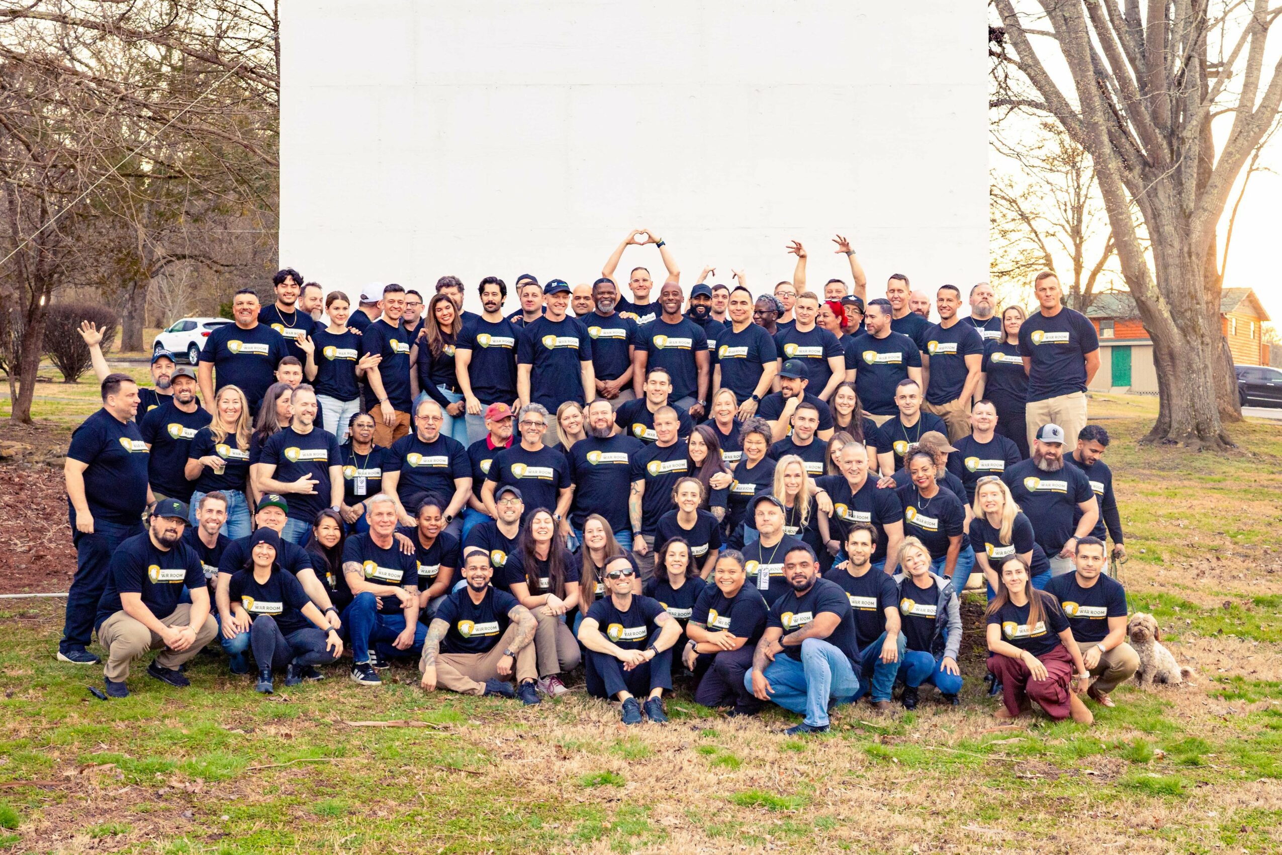 Group of people outdoors, standing and sitting, wearing matching dark shirts, with trees and a white wall in the background.